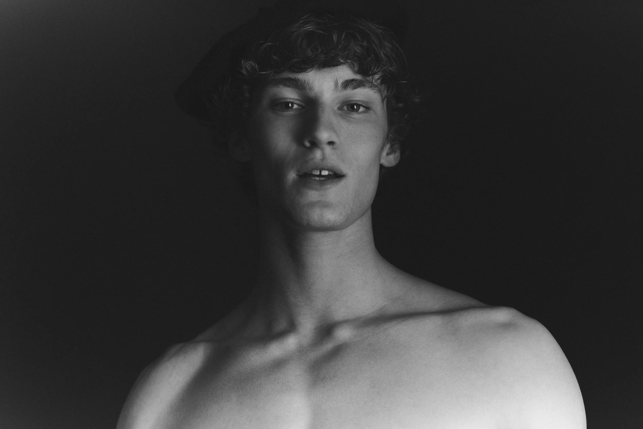 Black-and-white studio test portrait of a young man with curly hair, bare shoulders, lit against a dark background.