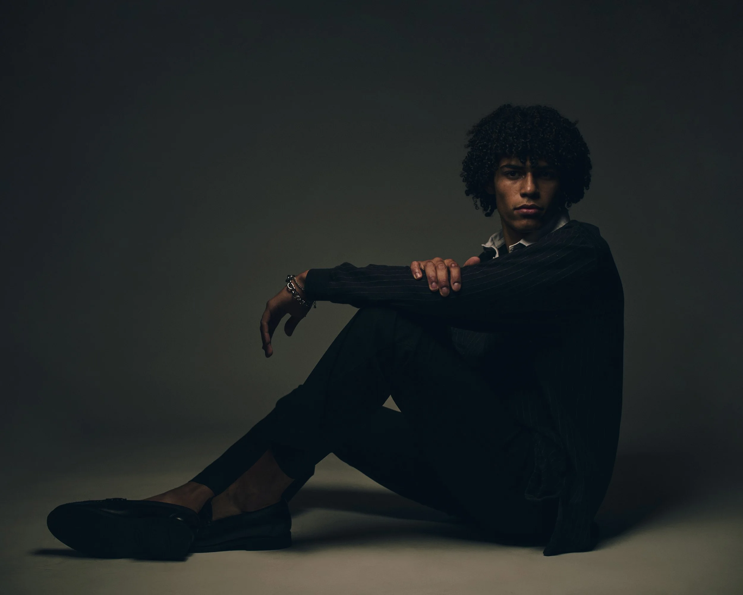 Low-key studio test portrait of a man with an afro, dressed in black and seated on the floor against a dark backdrop.