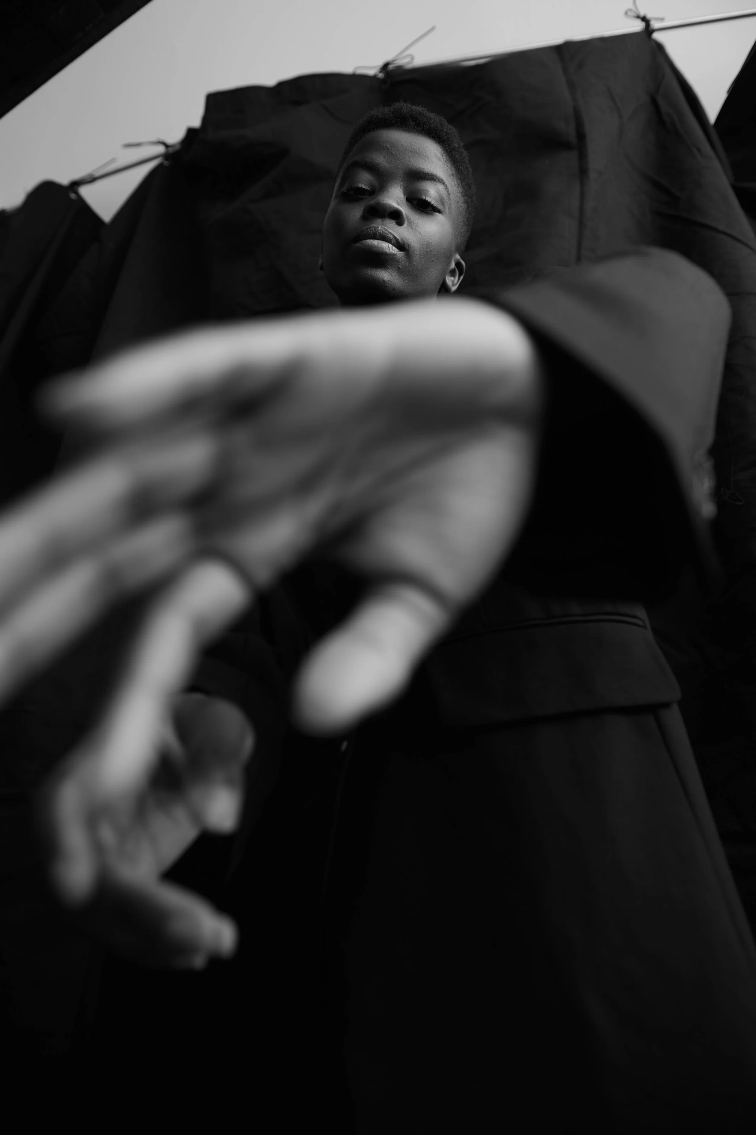 Black-and-white test shoot of a woman in a black tailored coat reaching a hand toward the camera, shot from a low angle.