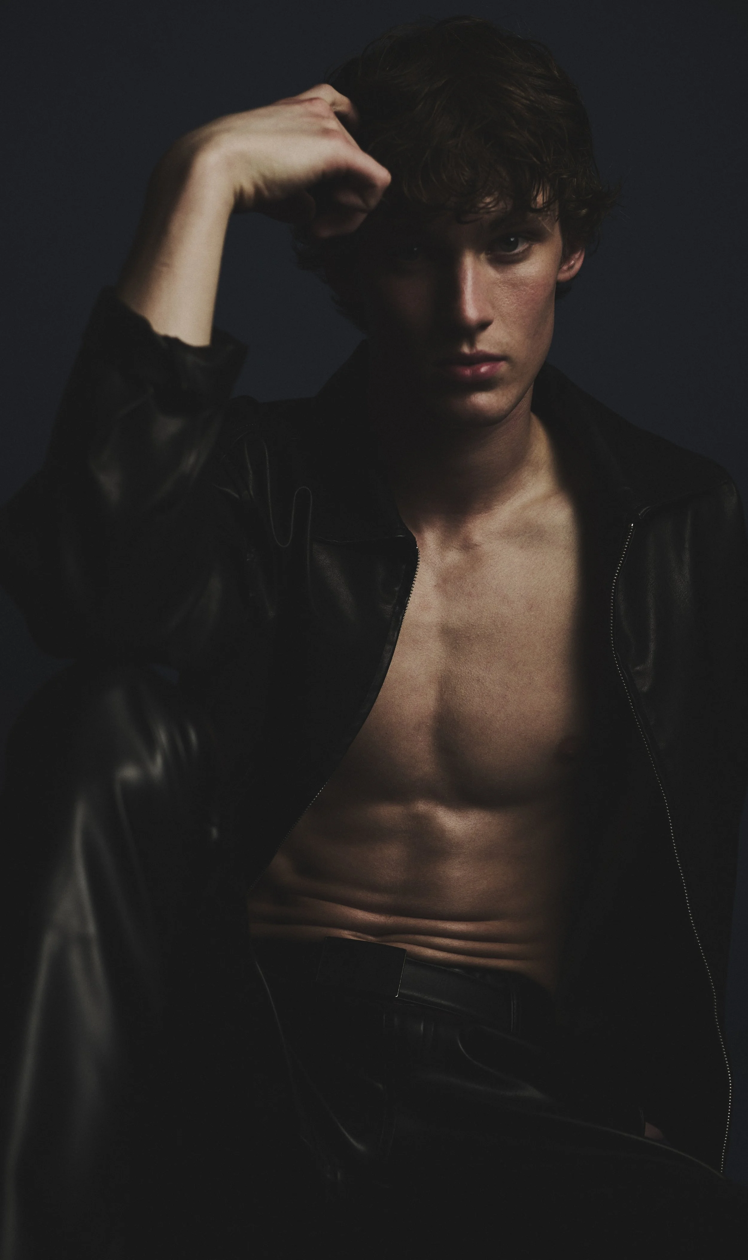 Low-key fashion test portrait of a young man in an open black leather jacket against a dark background.