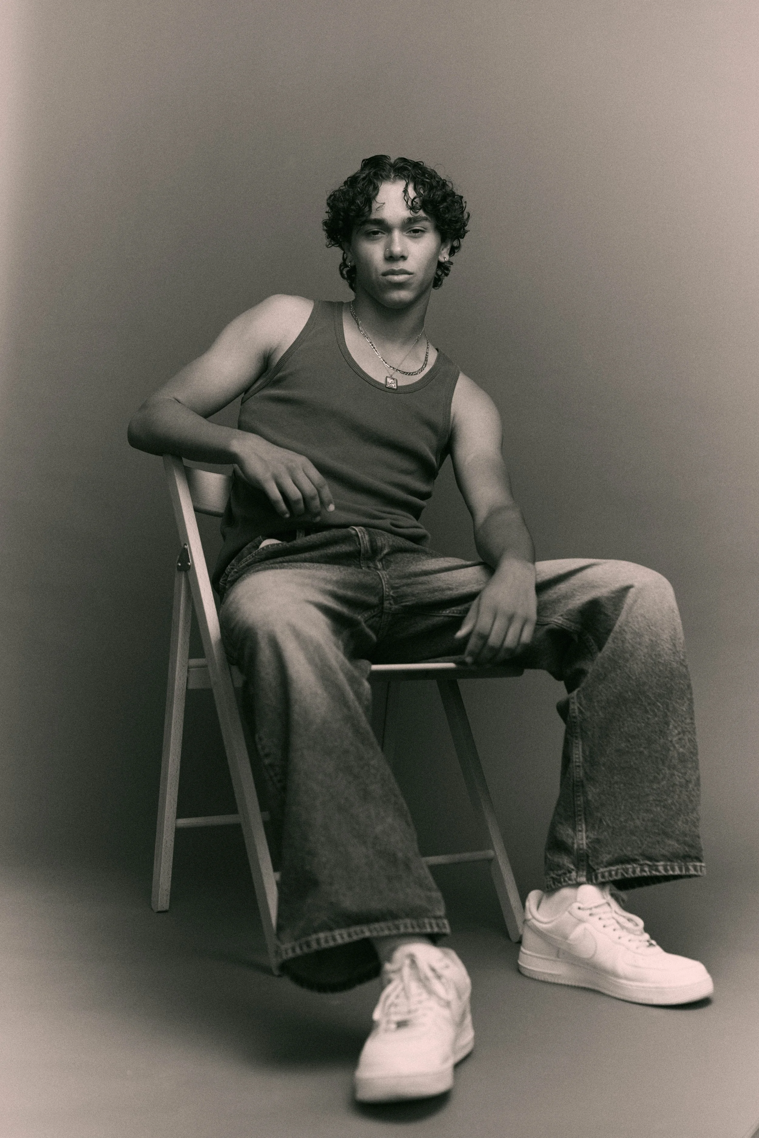 Studio test shoot of a man in a grey tank top, baggy jeans and white trainers seated on a stool, warm neutral backdrop.