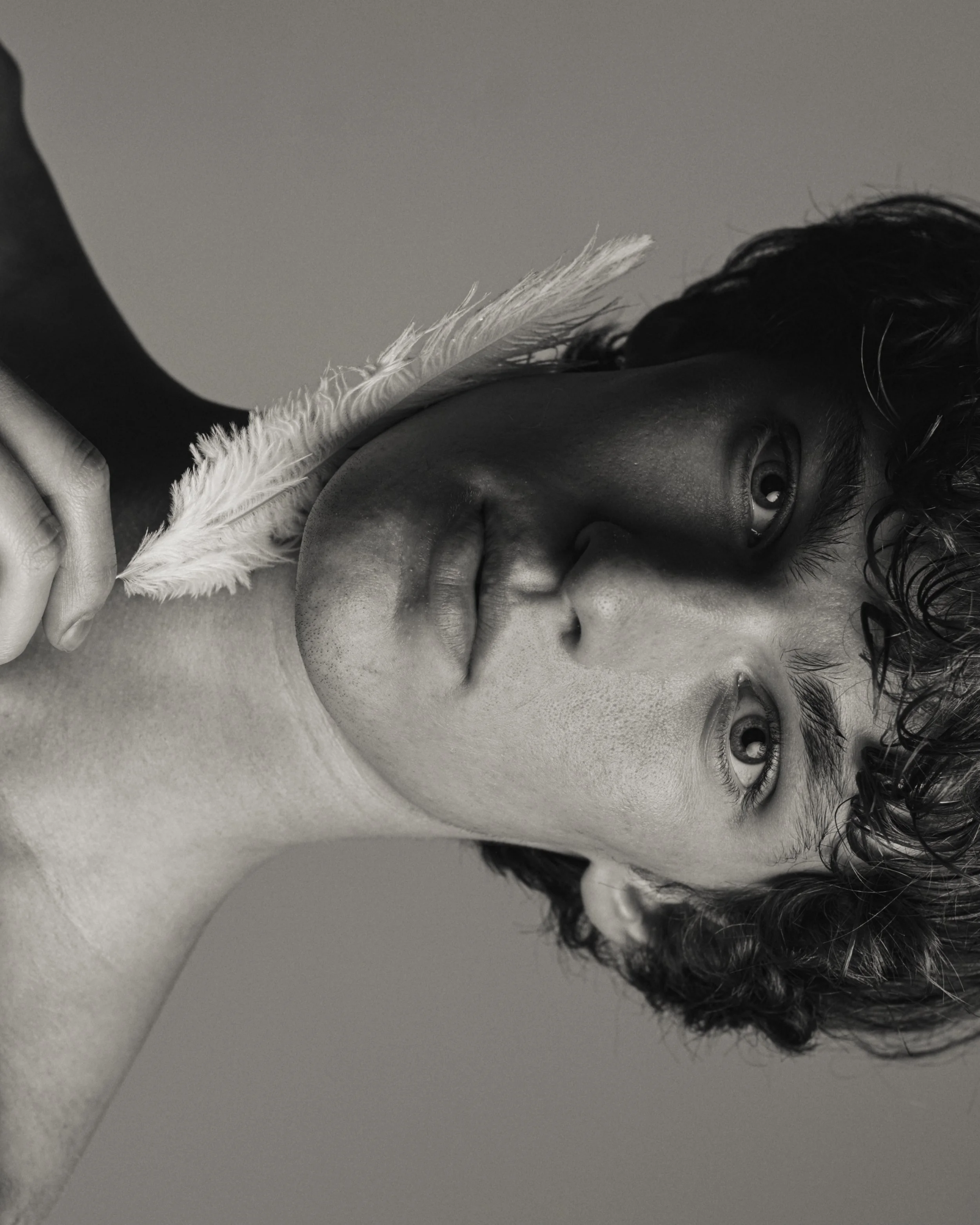 Black-and-white beauty test portrait of a man with curly hair lying on his side, a white feather held against his cheek.