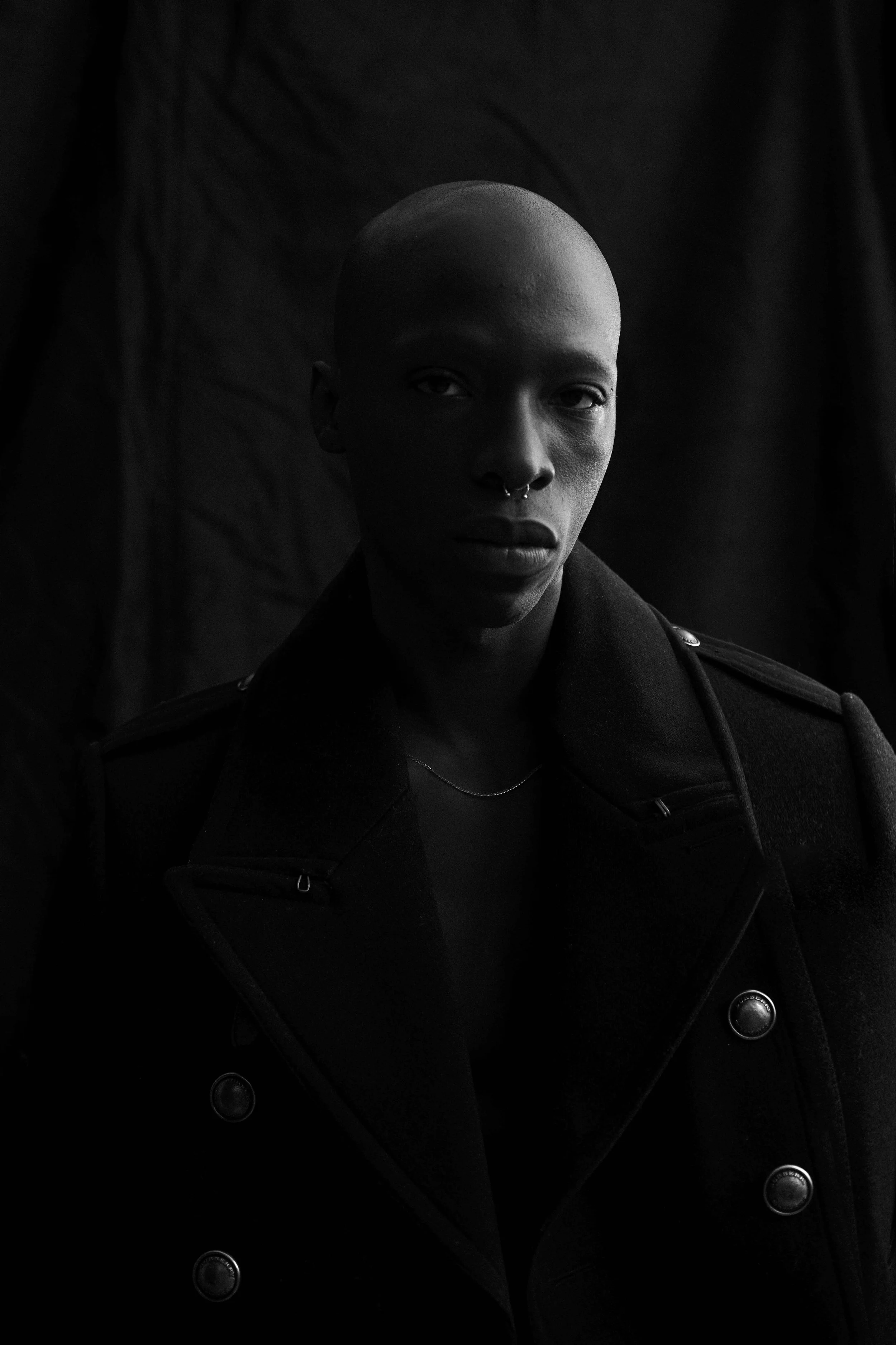 Low-key black-and-white test portrait of a man with a shaved head in a double-breasted coat against a black backdrop.