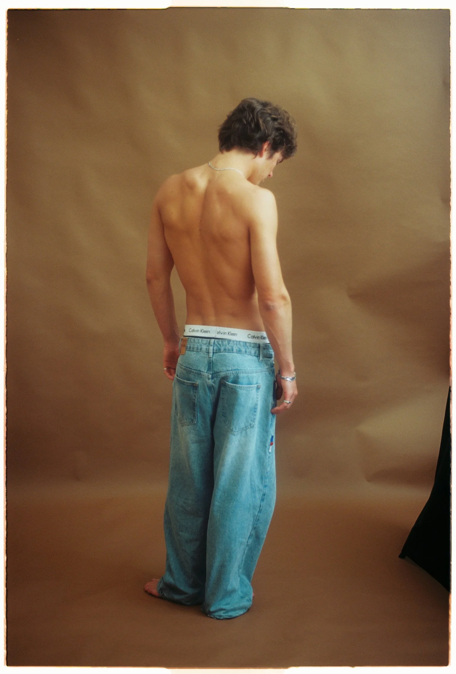 Studio test shoot of a shirtless man photographed from behind in loose blue jeans against a tan paper backdrop.