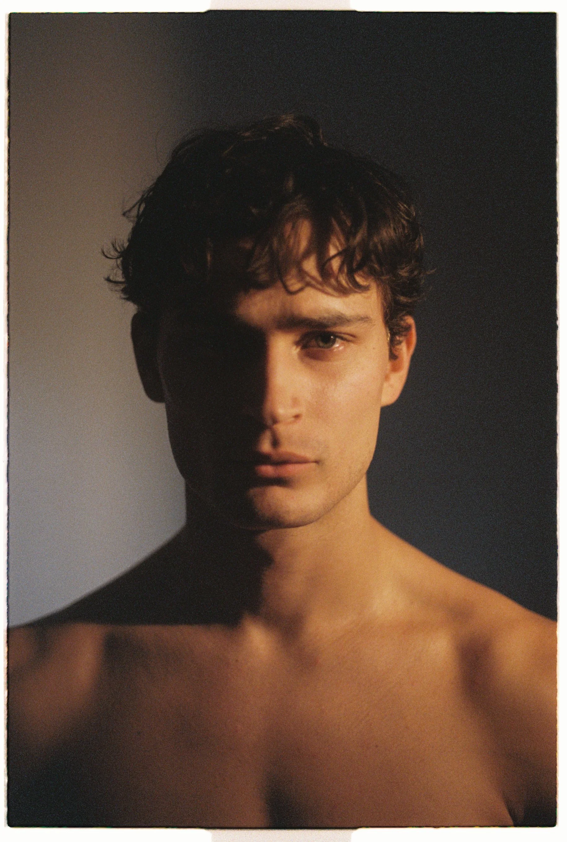 Warm studio test portrait of a young man with curly hair and bare shoulders, half his face in shadow.