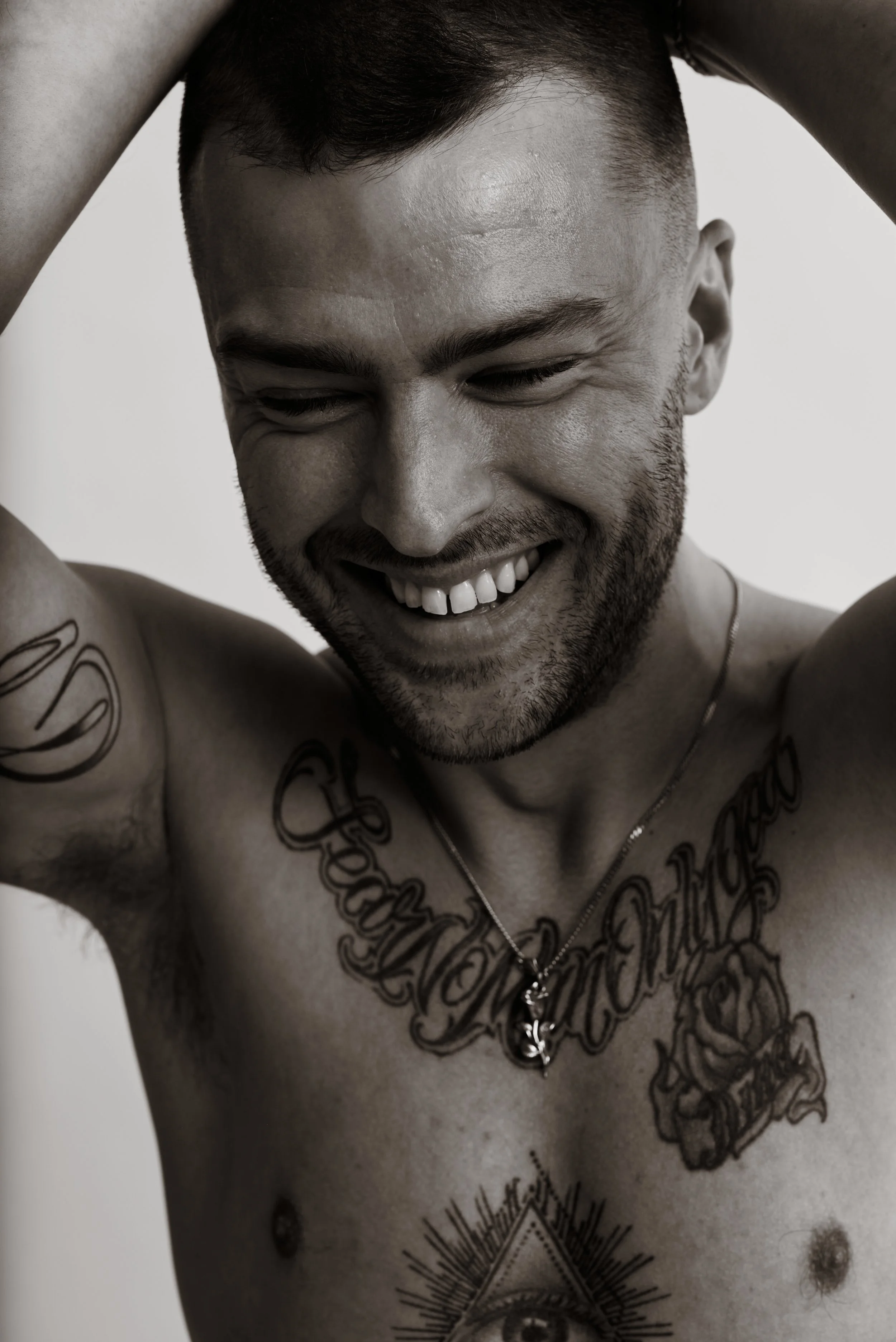 Black-and-white studio test portrait of a smiling tattooed man with his hands behind his head on a pale backdrop.