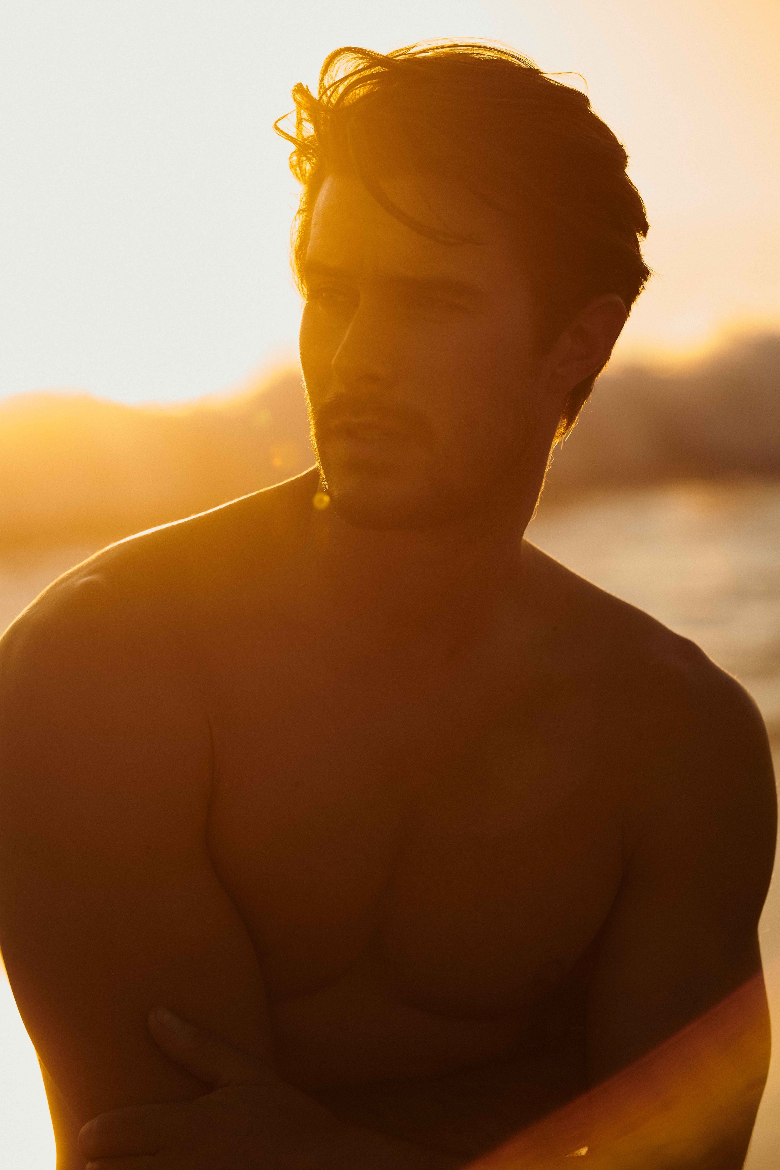 Backlit golden-hour test portrait of a shirtless man at the water's edge, sun flaring behind him.