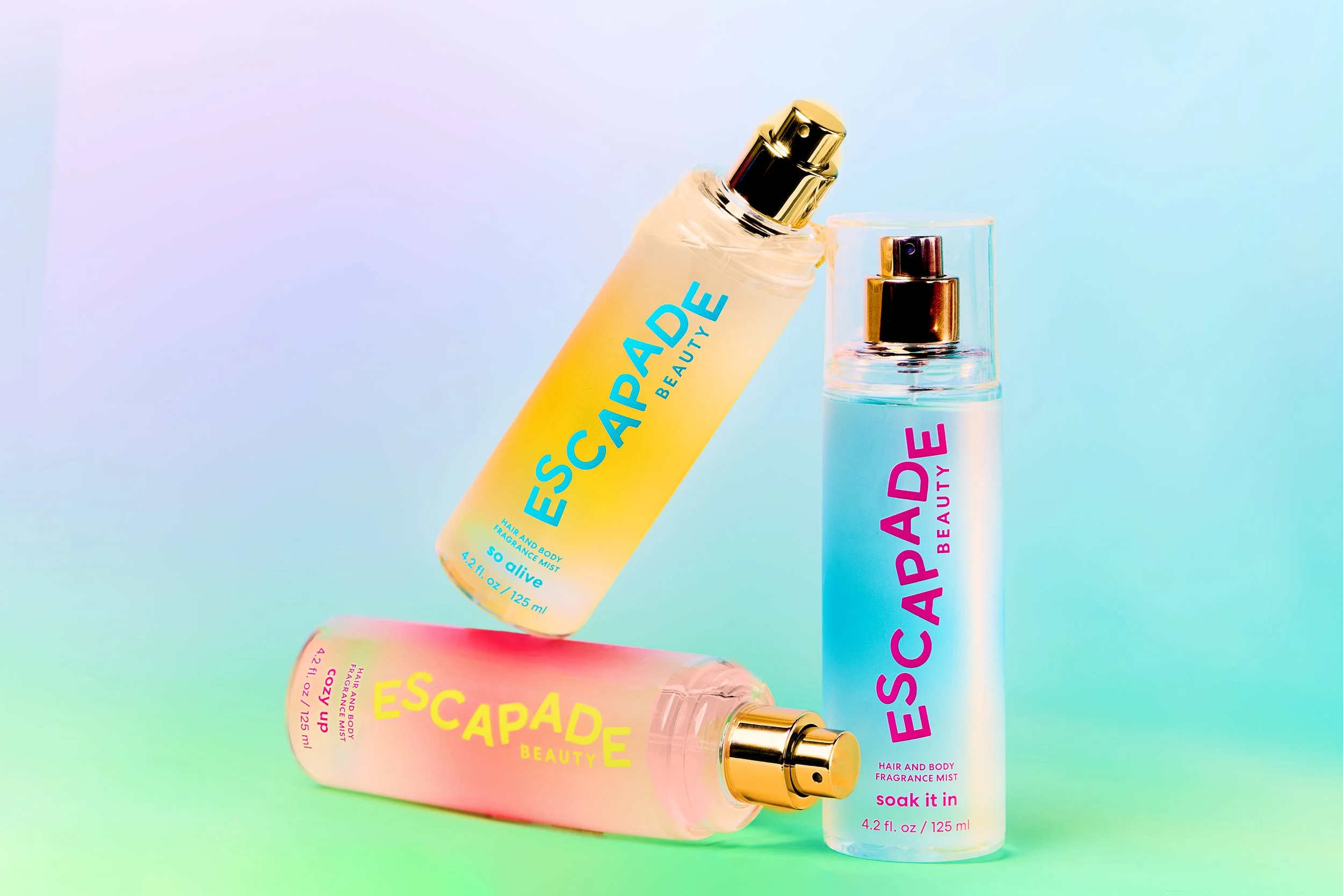 Colour-gradient product still life of three Escapade Beauty fragrance mists on a pastel set.