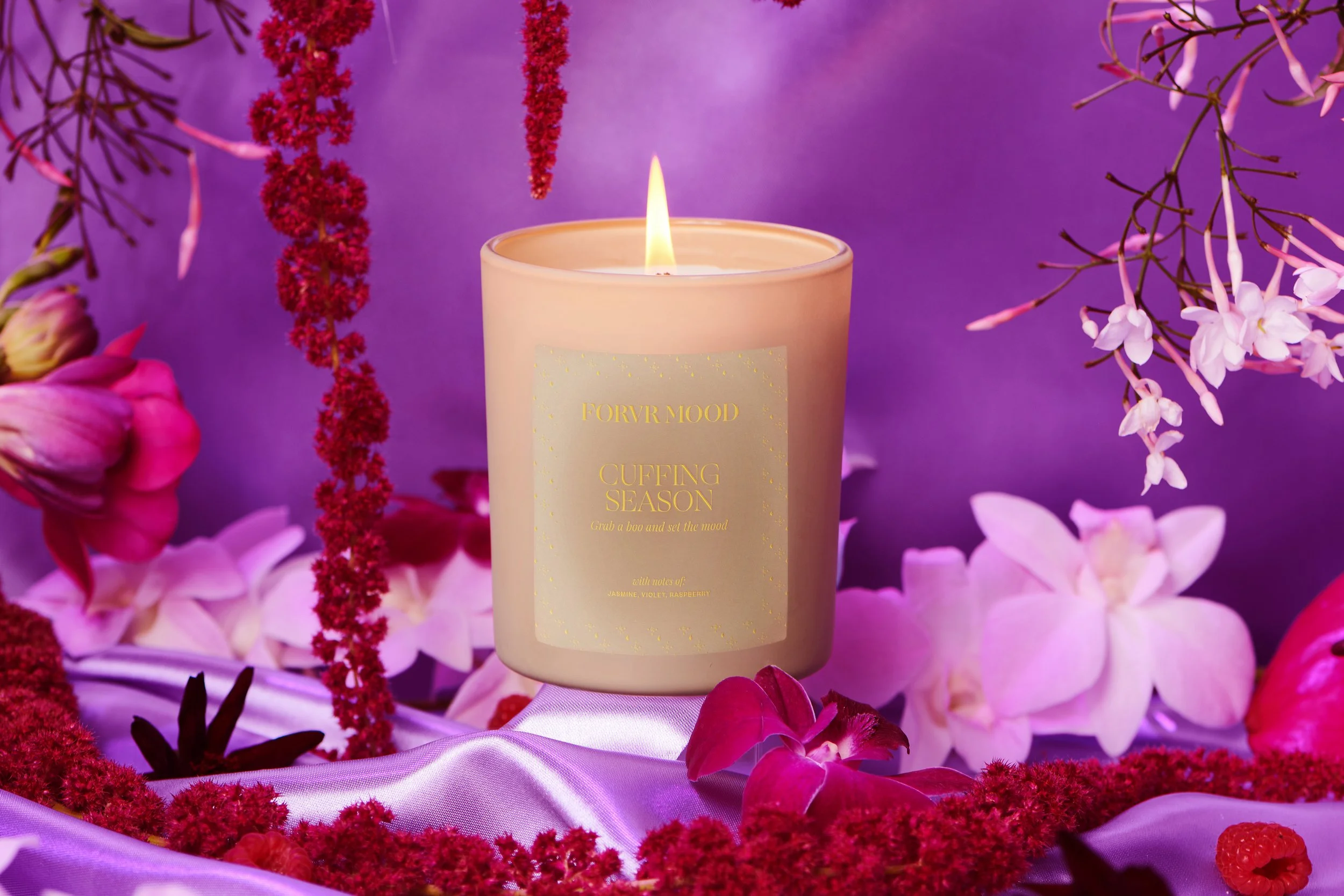 Built-set still life of the FORVR MOOD Cuffing Season candle lit among orchids and amaranth on purple satin.