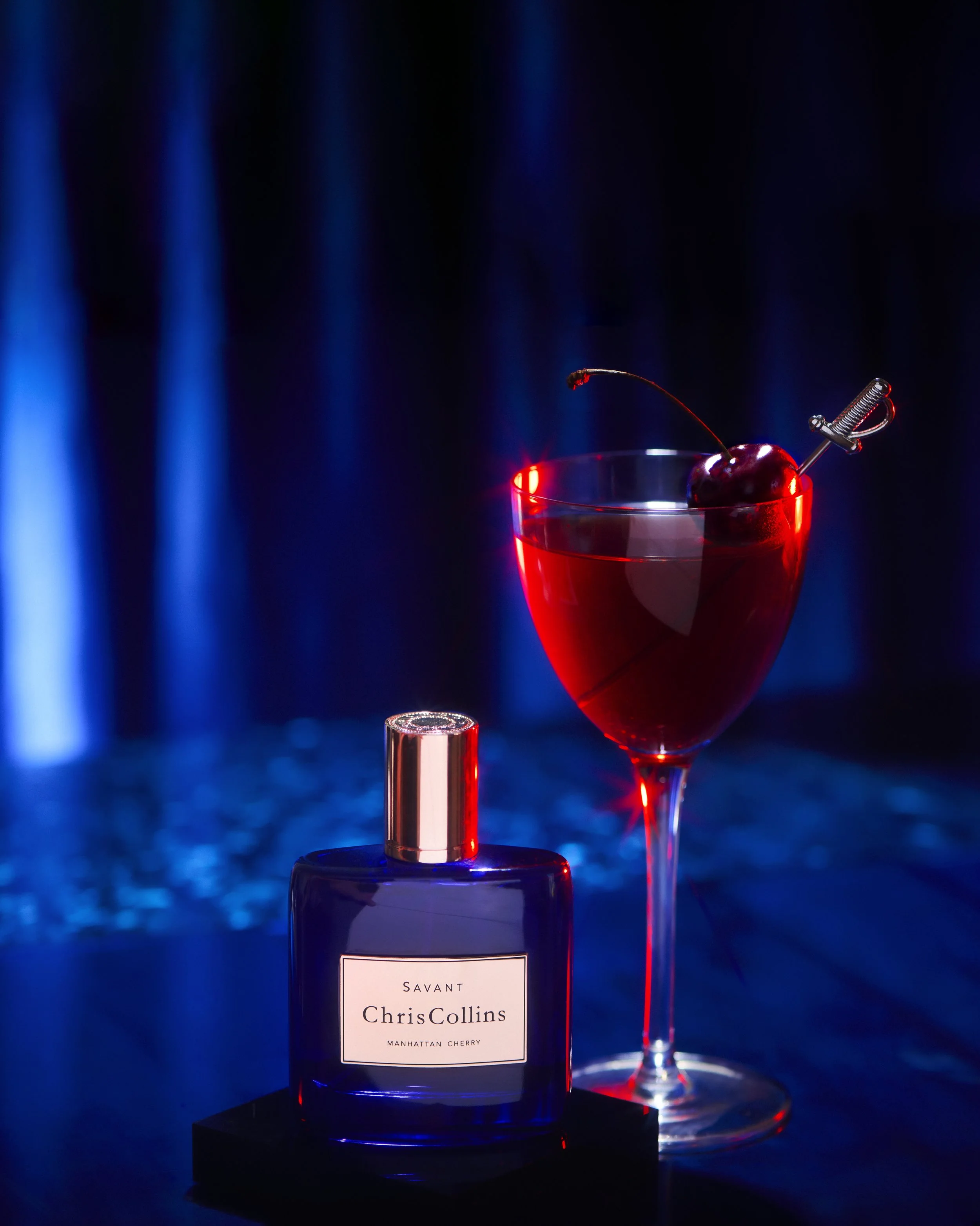 Fragrance still life of the Chris Collins Savant bottle beside a Manhattan cocktail under blue gel light.