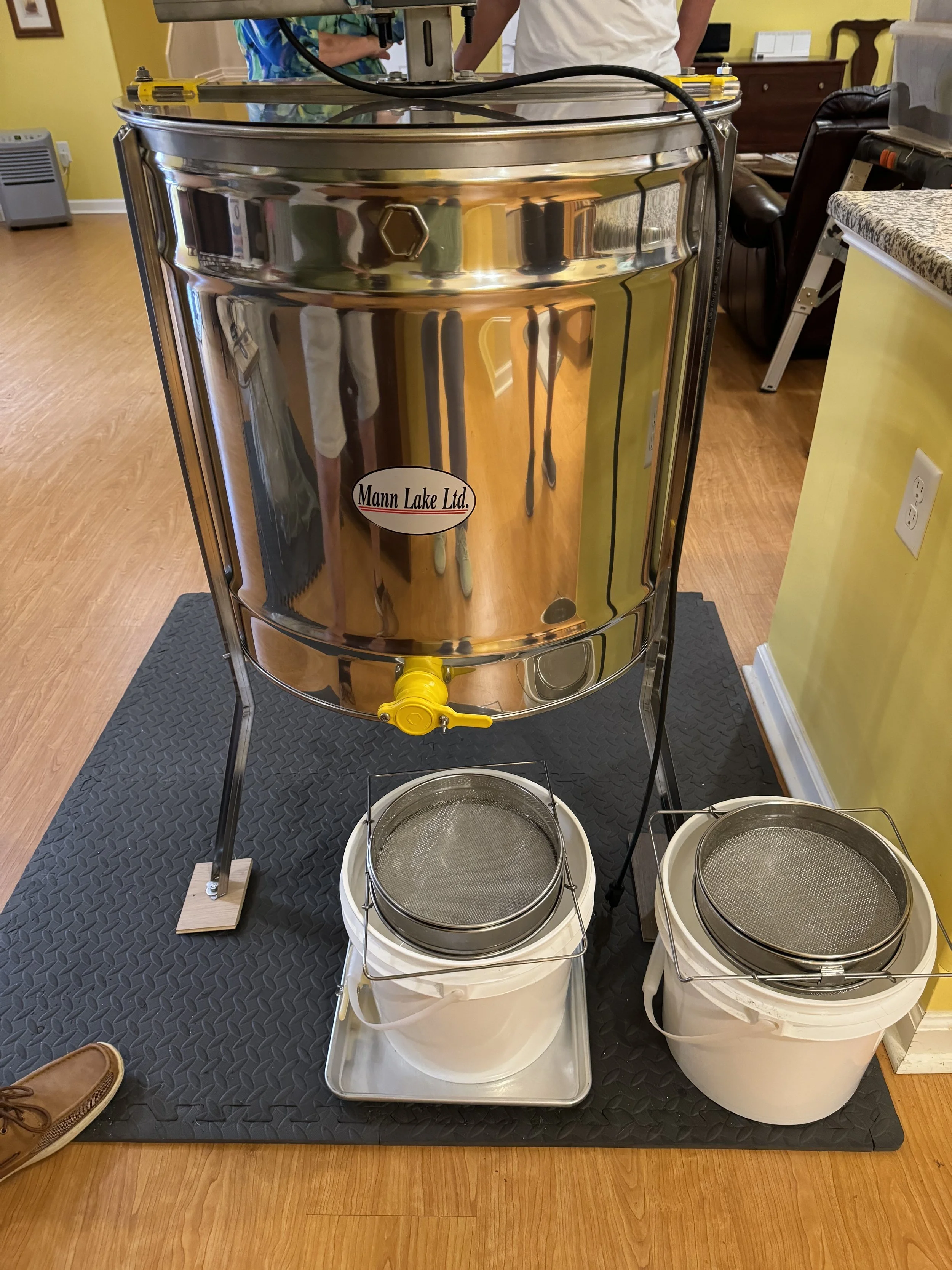 A sparkling metal honey extractor with a yellow spigot, sitting on a black rubber mat, with two white buckets with strainers underneath.