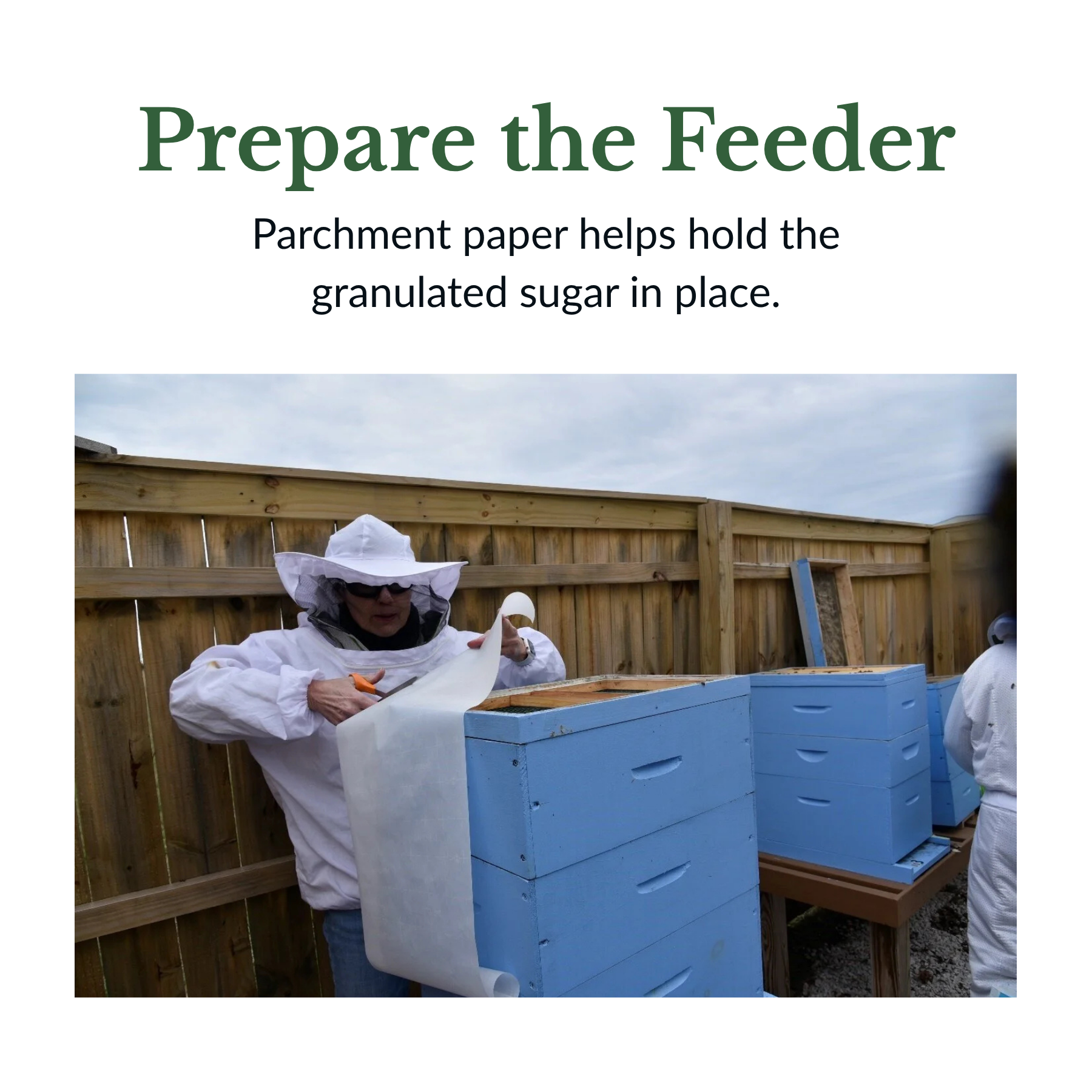 A person wearing a protective beekeeper suit and veil is handling a blue beehive box outdoors, with a wooden fence and cloudy sky in the background.