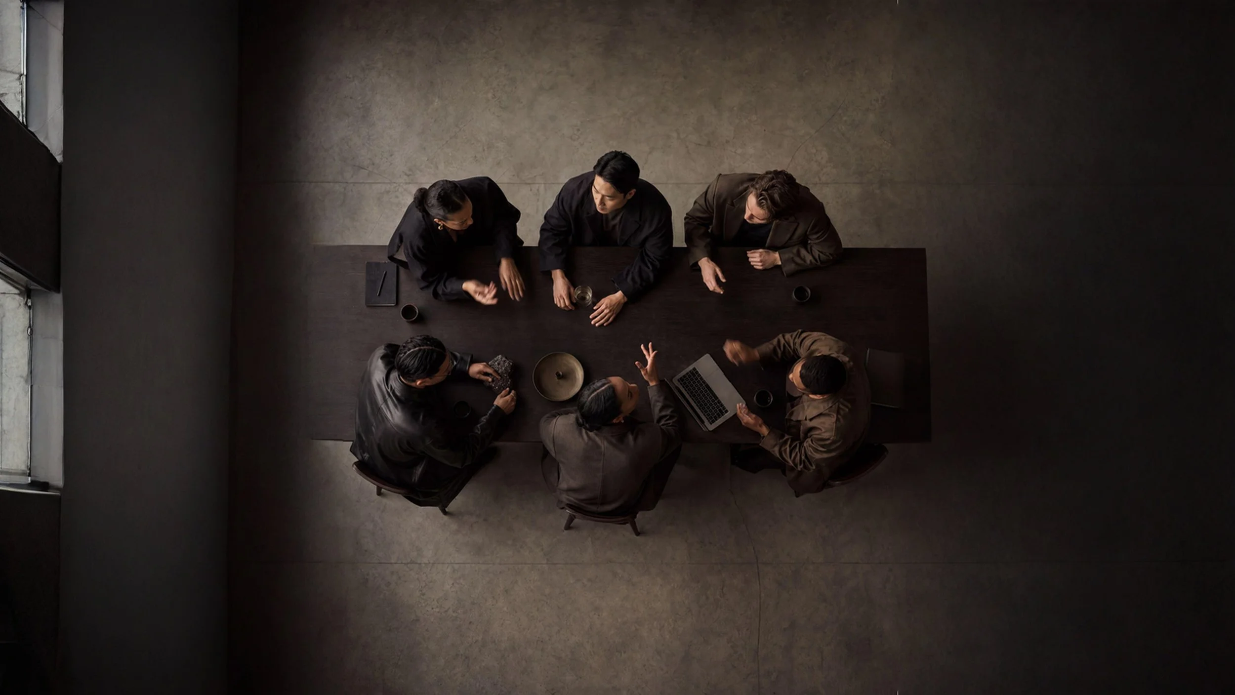 Top-down view of six people sitting at a dark rectangular table engaged in a discussion, with laptops, notebooks, and drinks on the table, in a modern, dimly-lit room.