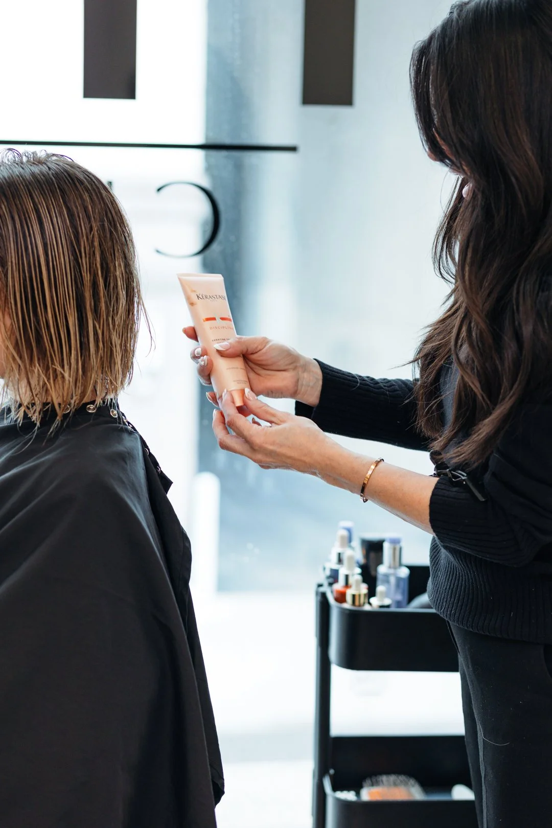 A hairstylist applying a hair product from a tube labeled 'Kérastase' on a client's wet hair in a salon.