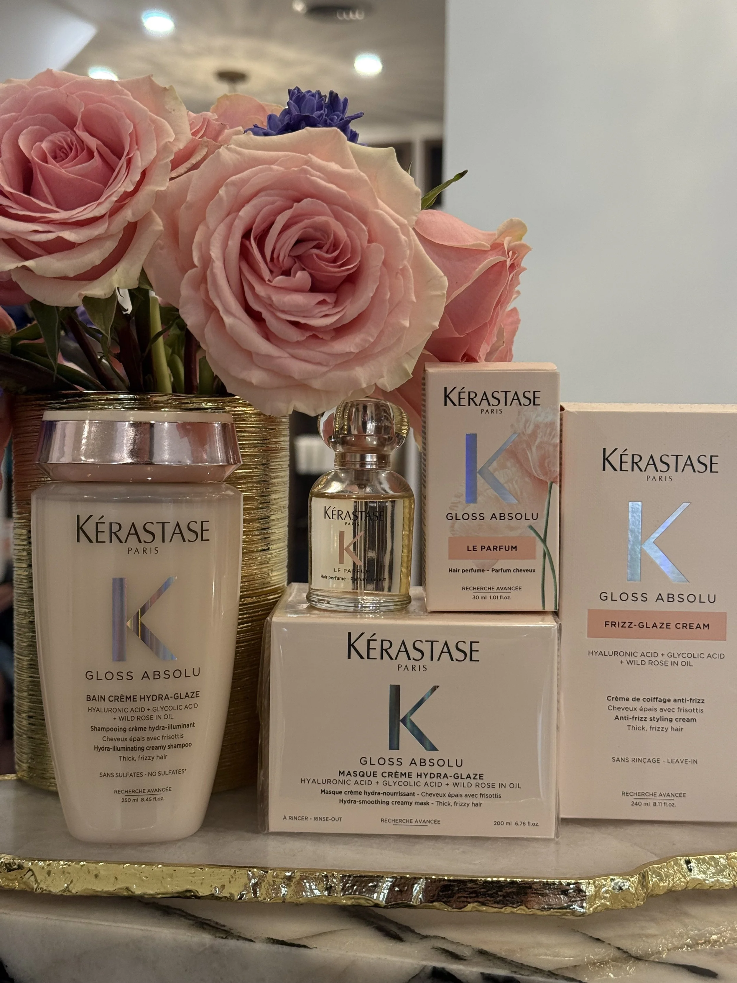A display of Kérastase hair care products with a pink floral arrangement, including shampoo, hair mask, and perfume bottles, on a gold tray.
