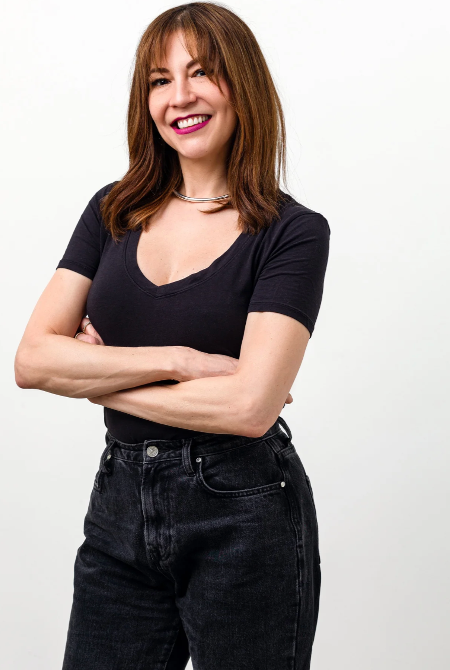 A woman with shoulder-length brown hair, smiling, standing with arms crossed, wearing a black t-shirt and black jeans, against a plain white background.