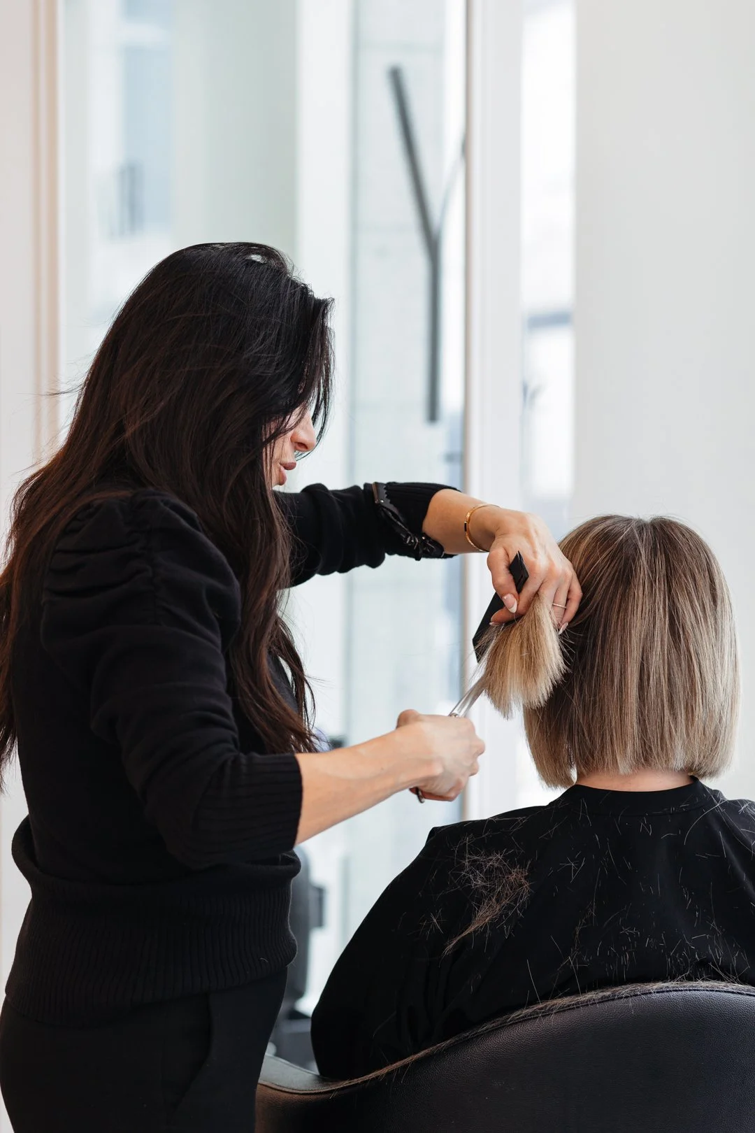 A hairdresser trims a woman's blonde bob haircut in a salon, with hair strands on her black cape.