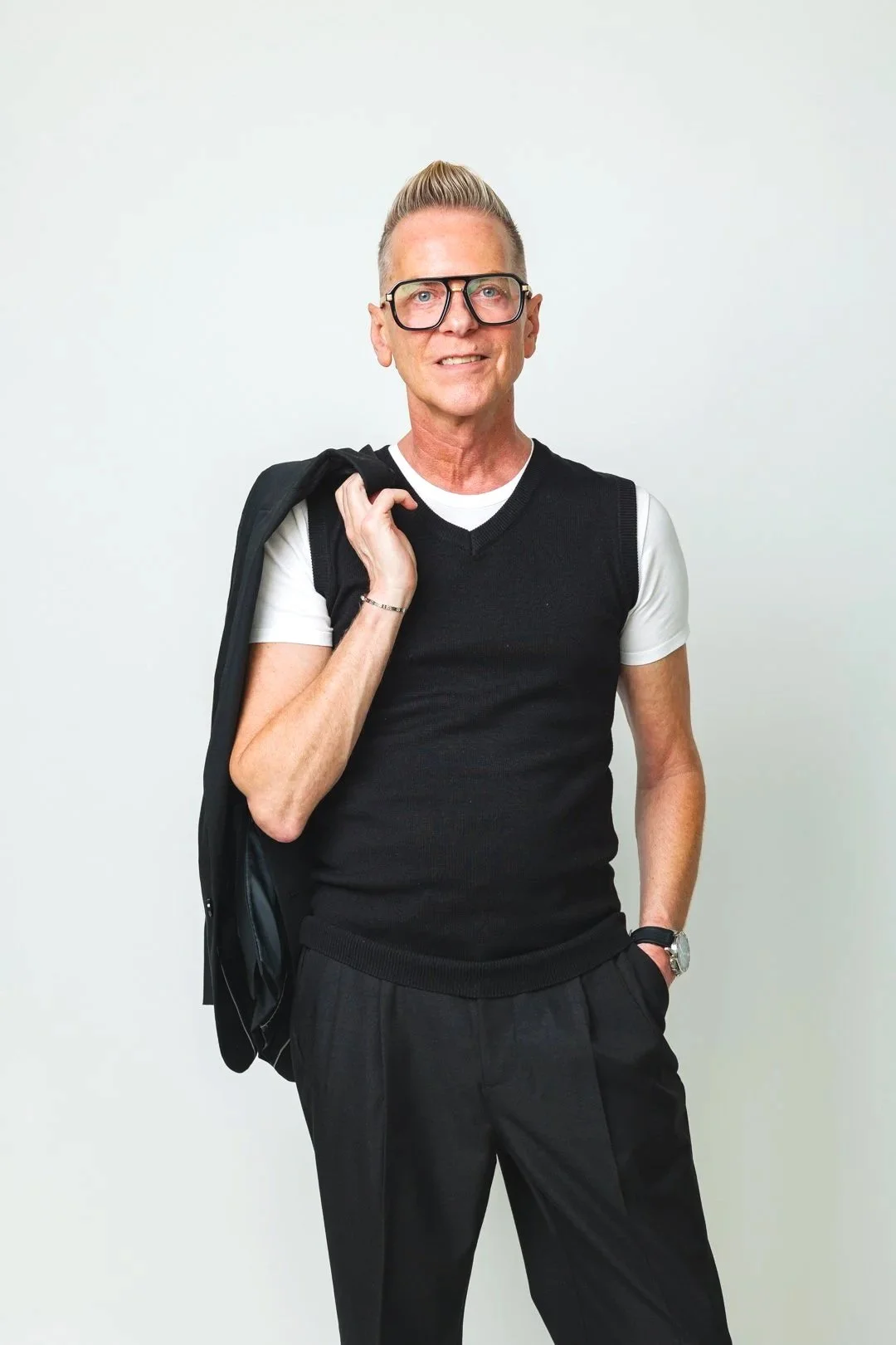 A man wearing black-rimmed glasses, a black and white plaid blazer with an embroidered crest, a white turtleneck, and black pants, standing indoors near a flower arrangement with pink roses.