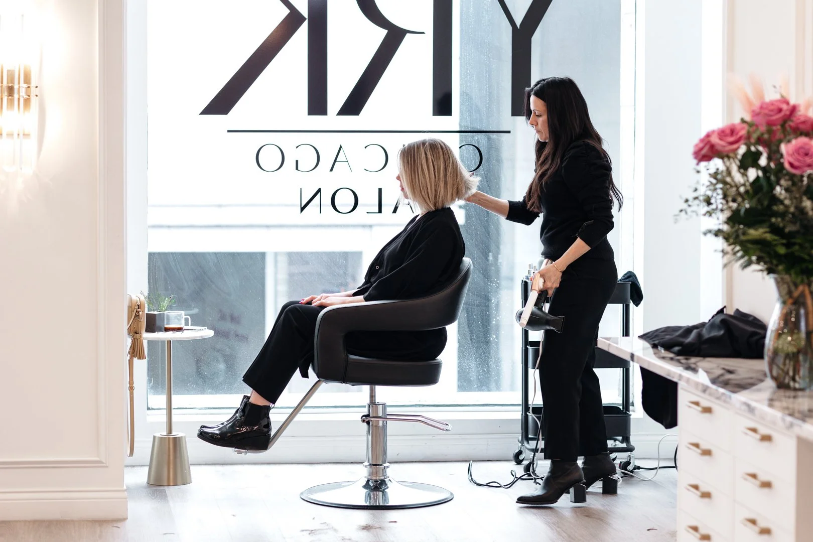 A woman sits in a black salon chair while a stylist, dressed in black, styles her hair. The salon features large windows, a marble counter with a vase of pink flowers, and a sign on the window that reads "ZRYO COACH salon".