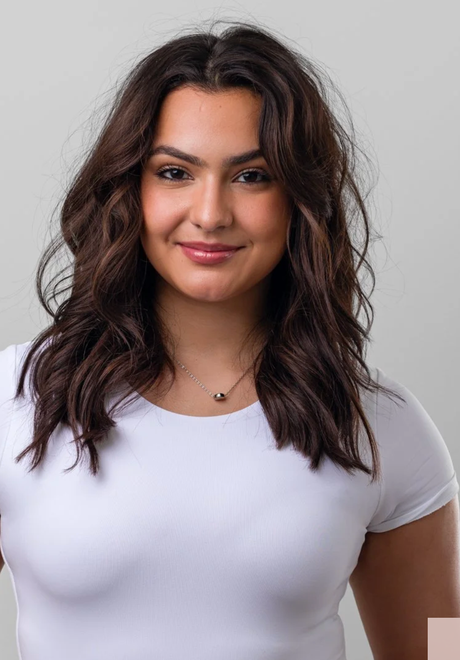 A young woman with wavy brown hair wearing a white shirt and a necklace, smiling at the camera against a plain gray background.