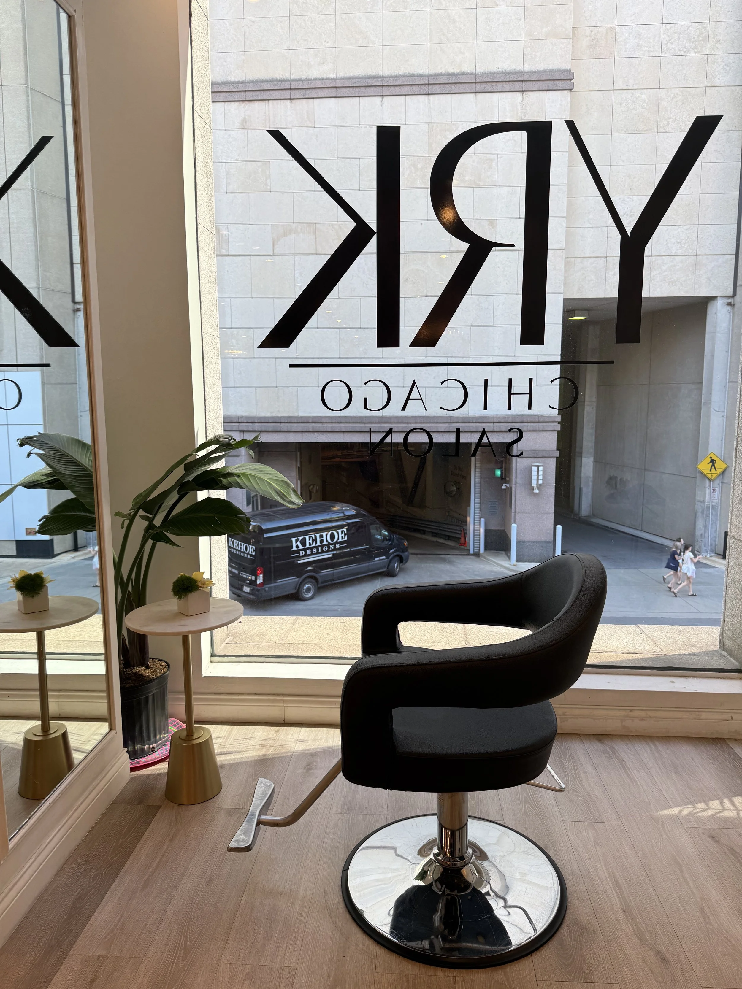 Interior view of a salon or spa with a black salon chair in the foreground, a small table with potted plants to the left, and large window with signage for 'KY' and 'CHIACCO SALON' on the glass. Outside, a black van with 'KEHOE DESIGNS' is parked, and pedestrians walk on the sidewalk.