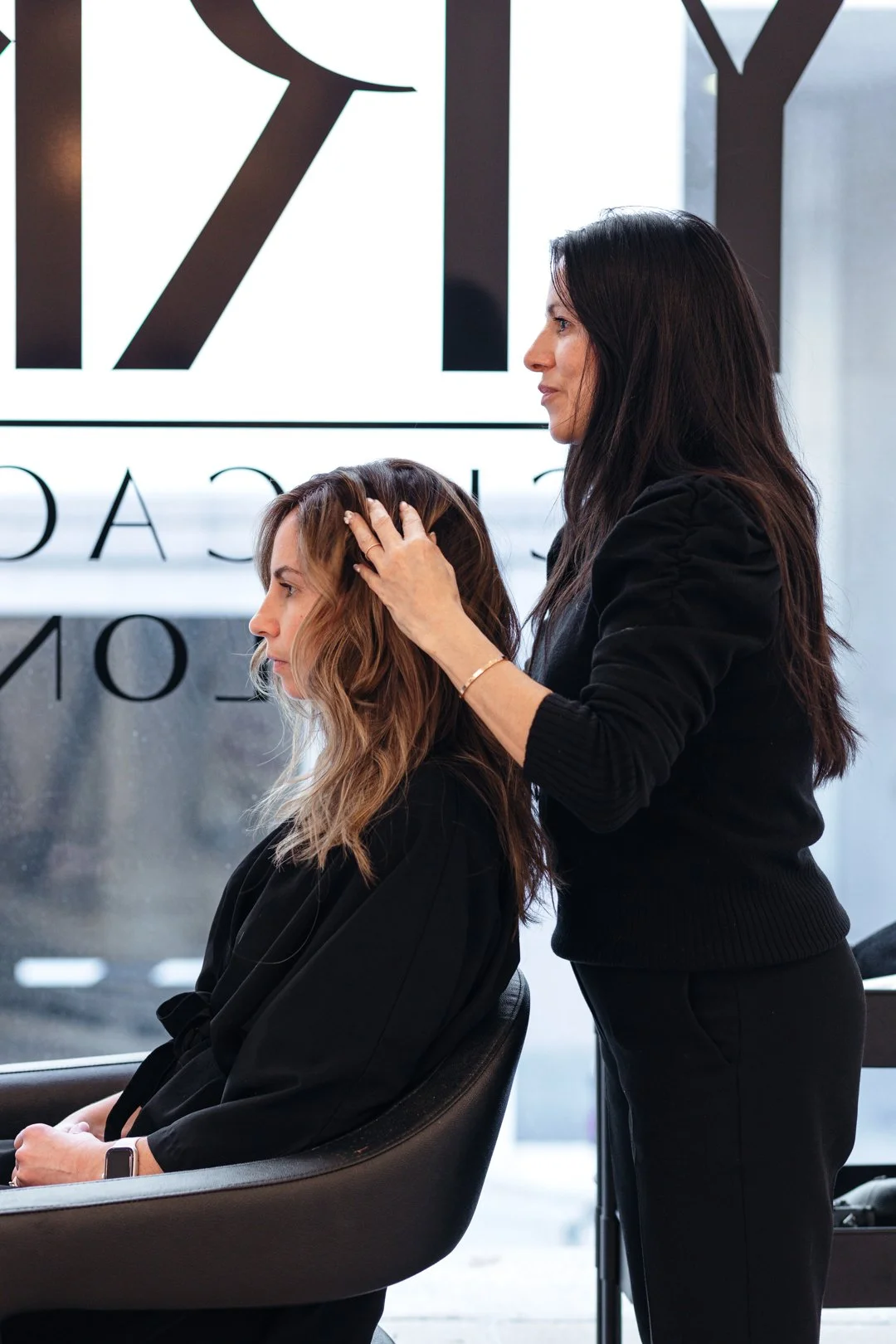 A woman getting her hair styled by a hairstylist in a salon with large window and logo in background.