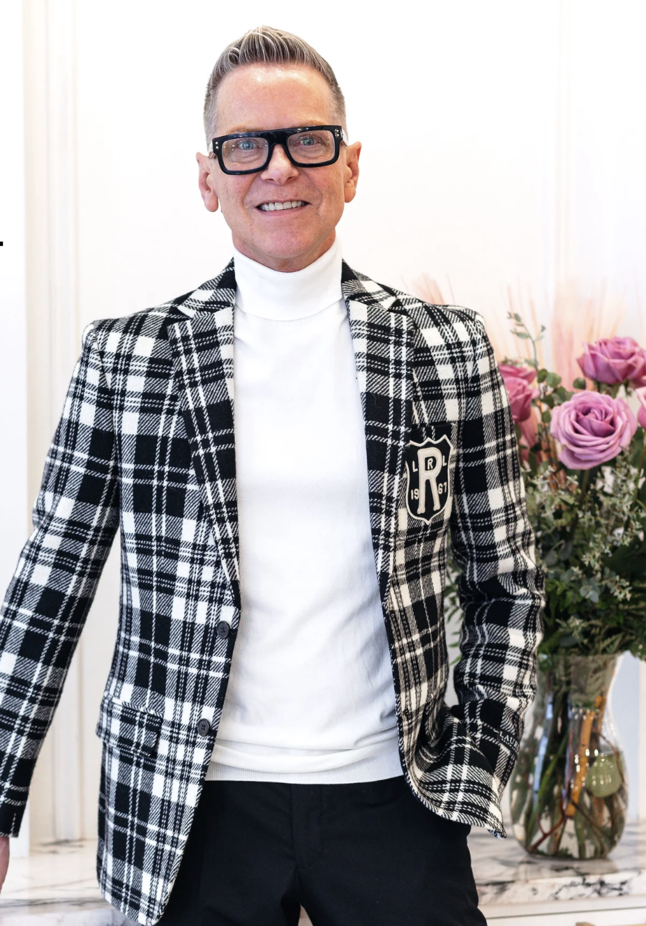 Man wearing black glasses, a black and white plaid blazer with a crest on the left pocket, a white turtleneck, and black pants, standing indoors with pink roses in the background.
