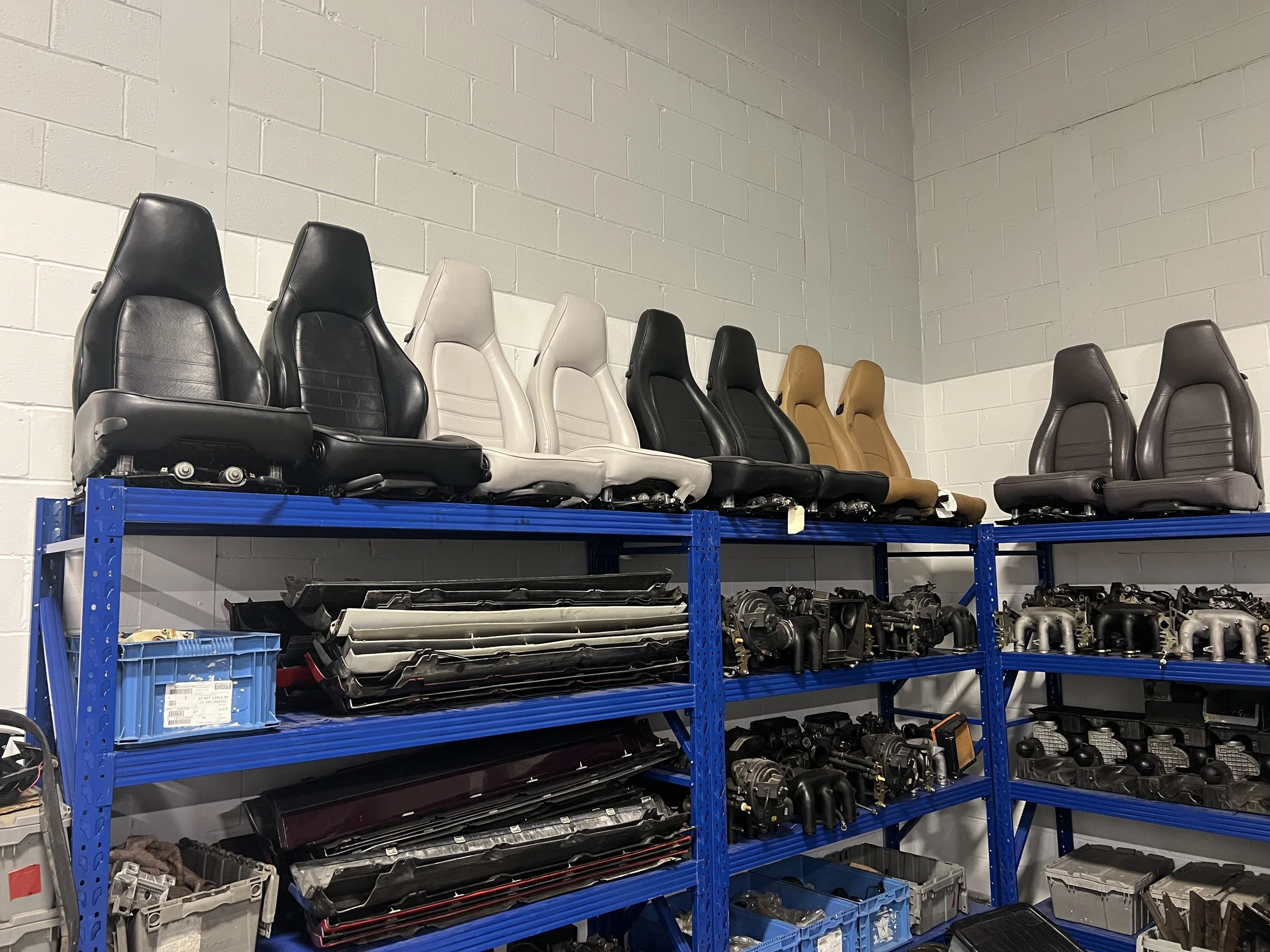 A blue metal shelving unit holding car seats, car parts, and storage bins in an industrial setting with cinder block walls.