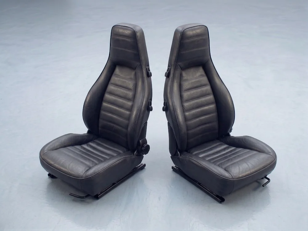Two black leather car seats with high backs and horizontal stitching, placed on a plain light gray floor.
