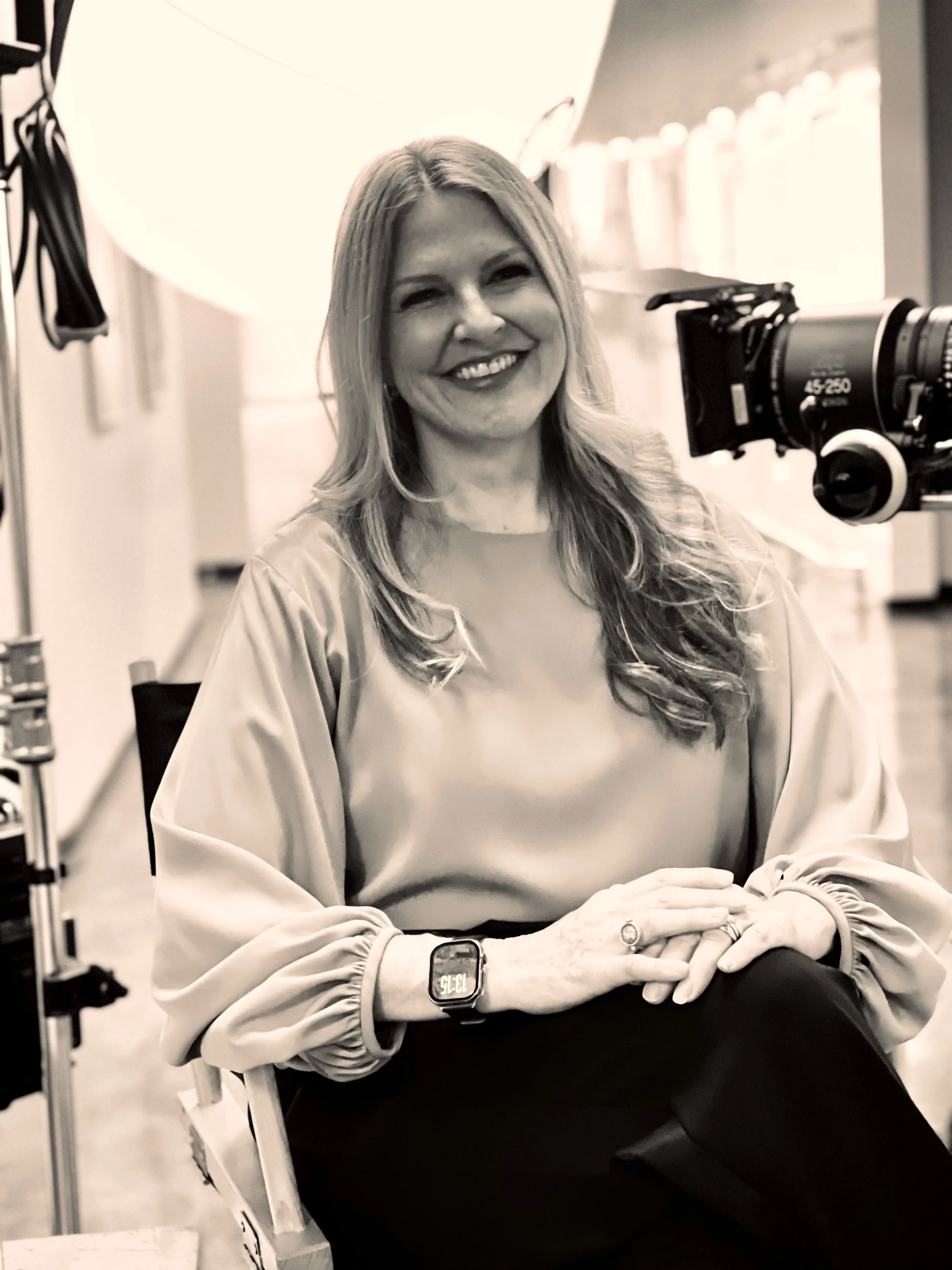 Black and white photo of a smiling woman sitting with her hands crossed on her lap, near professional photography equipment and a large light, in an indoor studio setting.