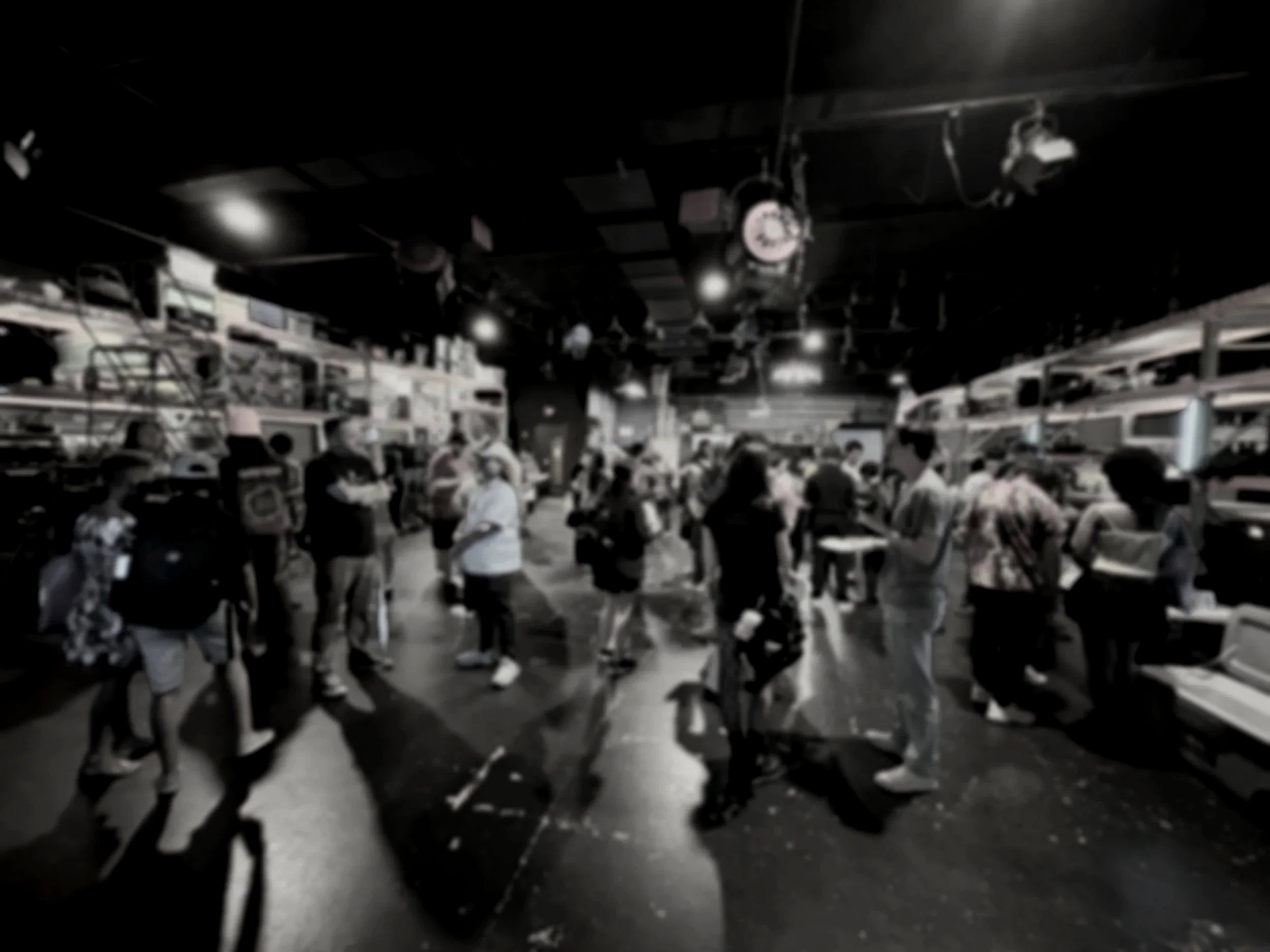 Black and white photo of people walking and socializing in an indoor space with a ceiling decorated with hanging lights and a clock.