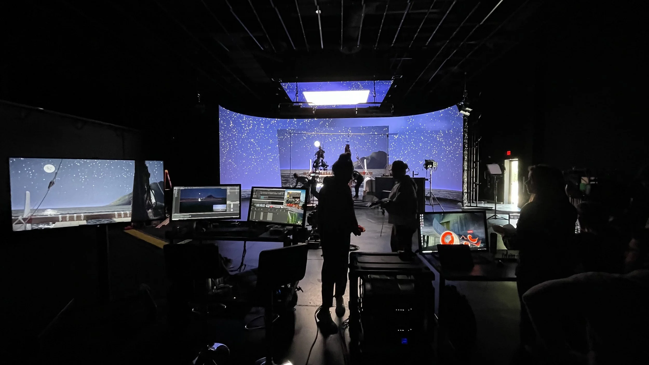 A behind-the-scenes view of a stage production being filmed, with crew members working at computer monitors and operating equipment, and a set featuring a night sky with stars.