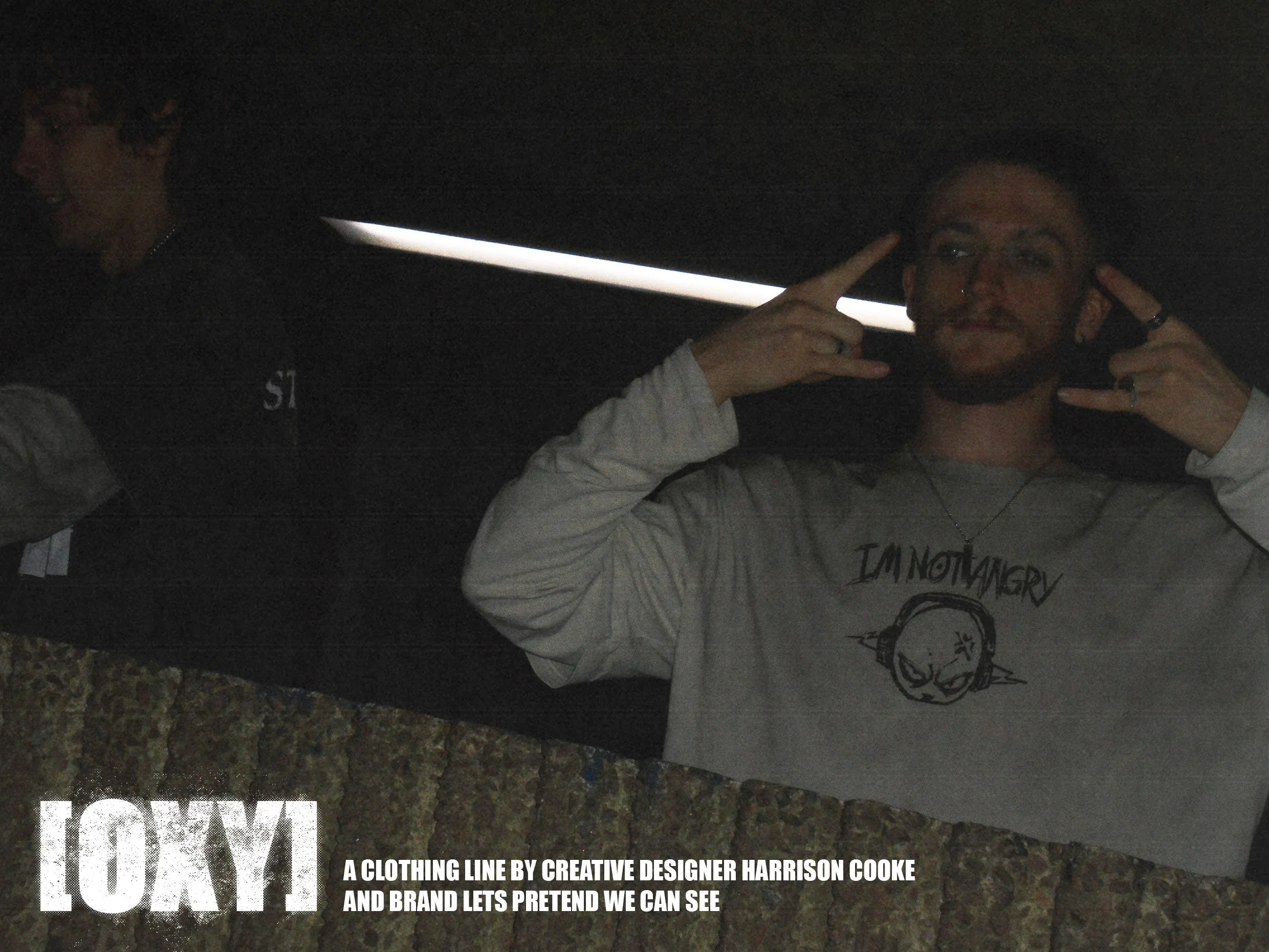 Two people in a dark environment, one person with glasses and a nose ring, wearing a gray shirt, making a hand gesture, with a partial view of another person with purple hair in the background. There is text at the bottom promoting a clothing line by