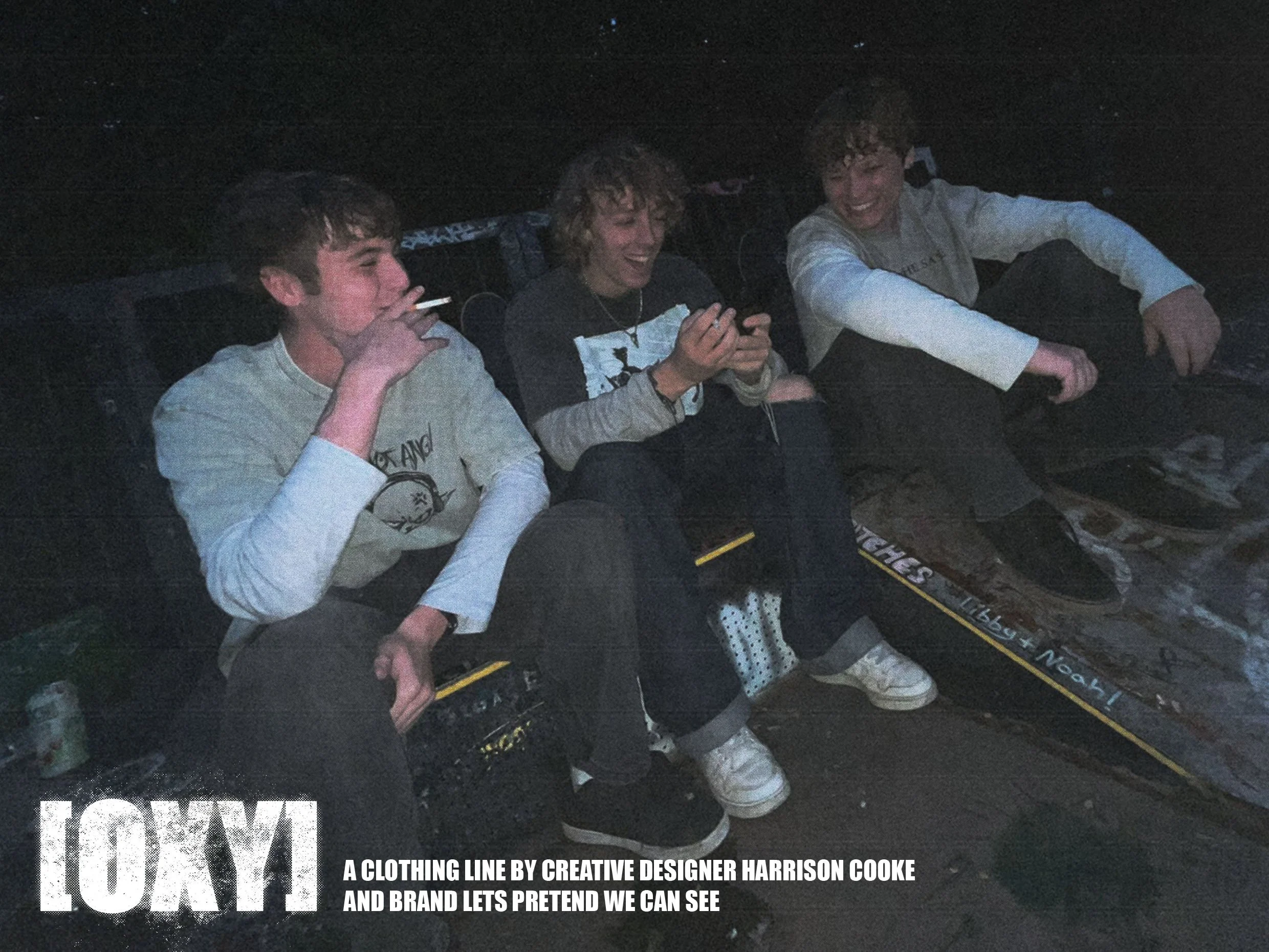 Four young boys sitting on the ground outdoors at night, smiling and laughing, with one smoking a cigarette, and wearing casual clothing.