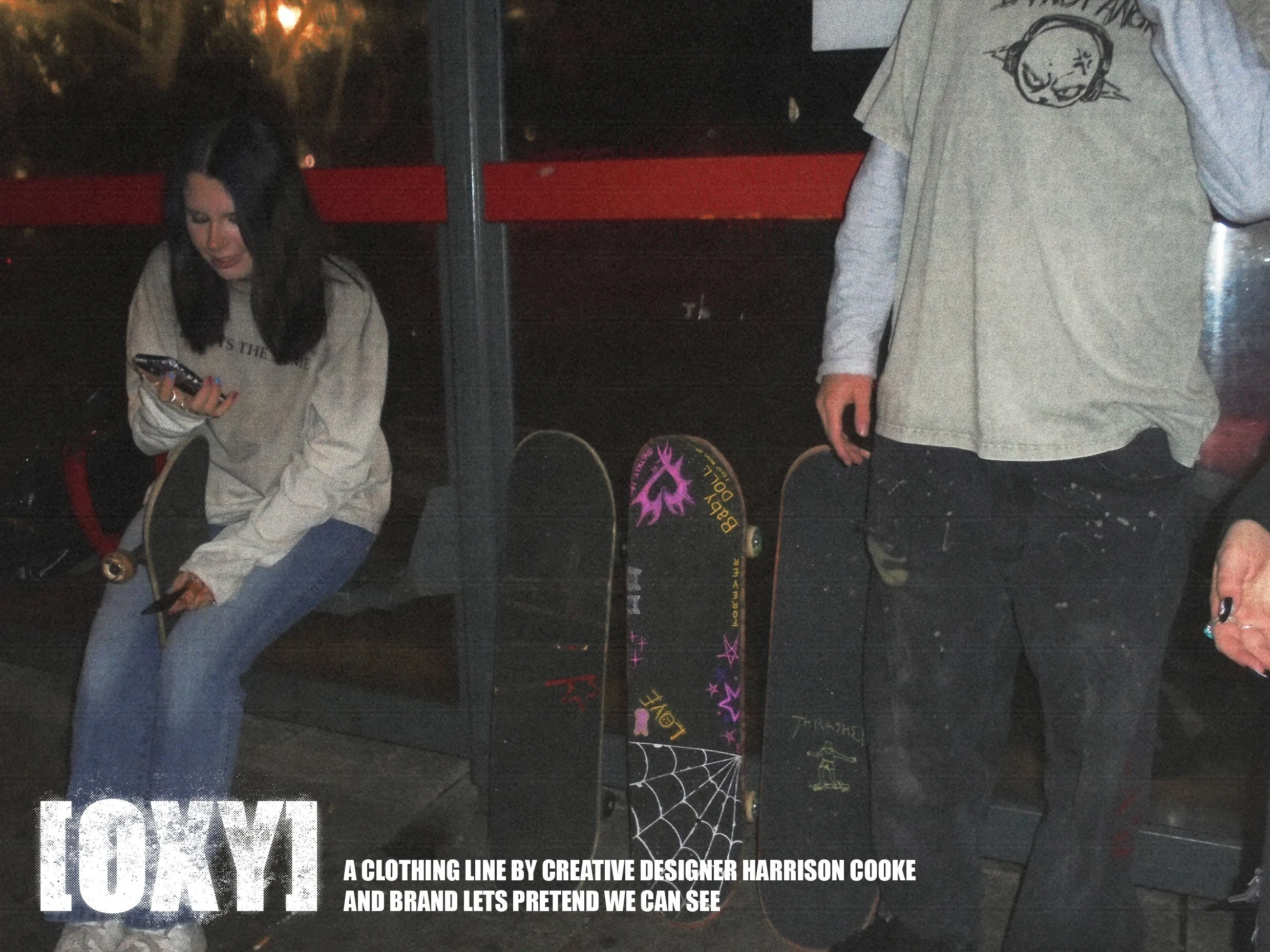 A woman with black hair and light skin is sitting while looking at her phone. To her right, a person with light skin, wearing a beige hoodie and black pants, is standing and reaching for a skateboard with colorful graphics, including spiderweb and st