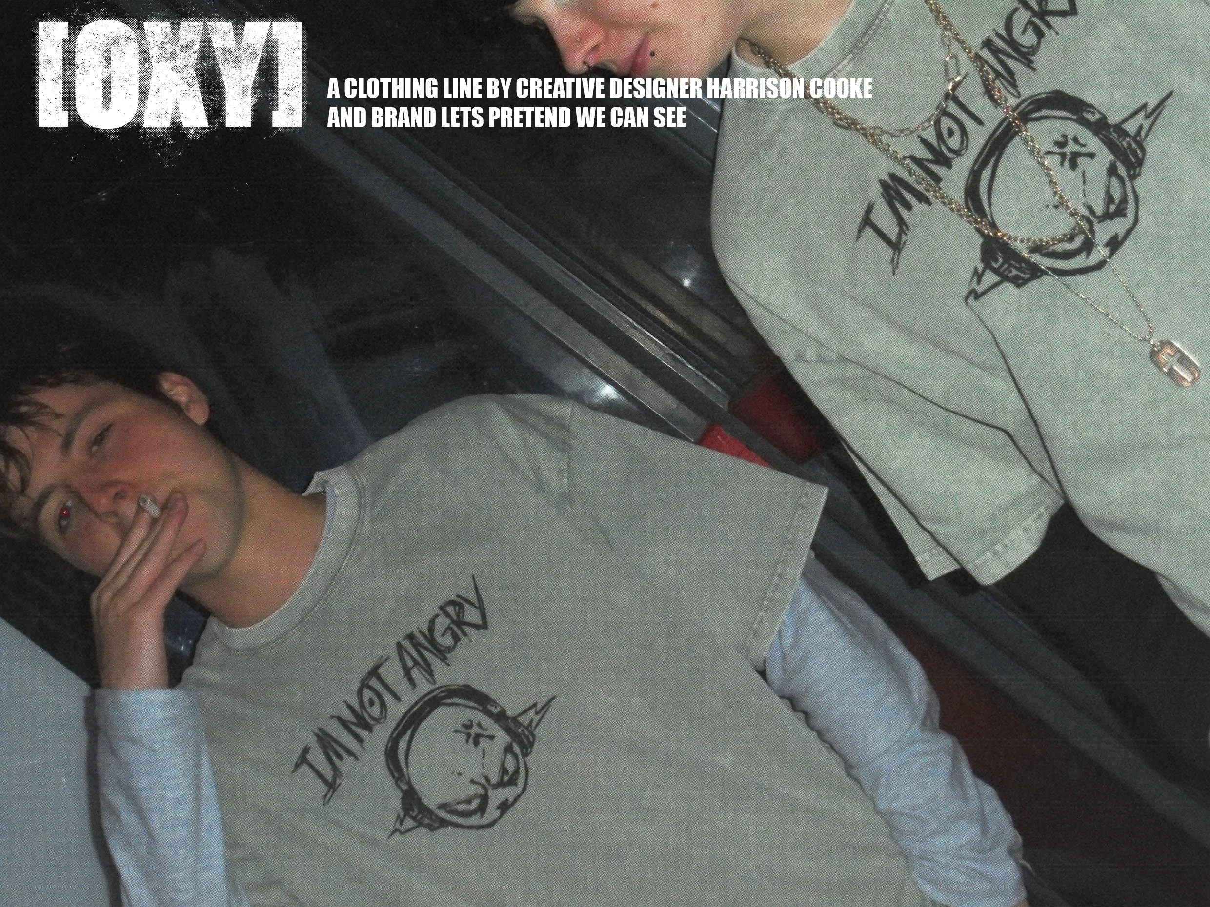 A person with short dark hair and piercings on their face, wearing a gray graphic t-shirt with a watch and layered necklaces, sitting at a table with a white T-shirt featuring a logo and text.