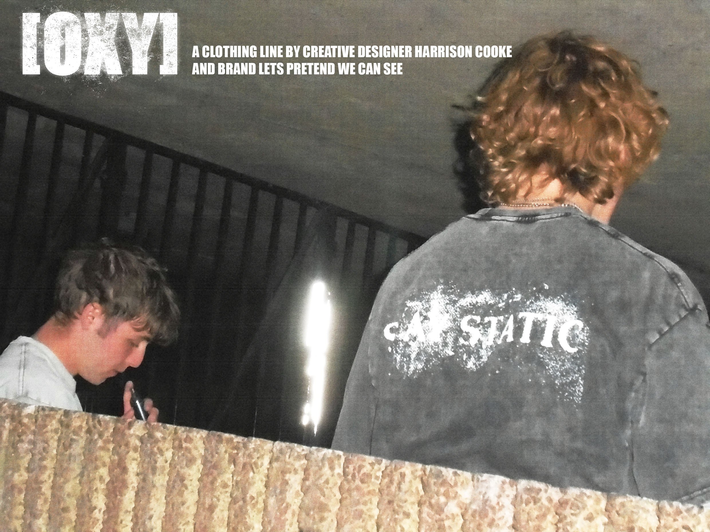 Back of a person with curly hair wearing a dark T-shirt with partially visible white text and a woman with short hair looking down at her phone, standing in front of a carpeted staircase with black railings and a granite counter.