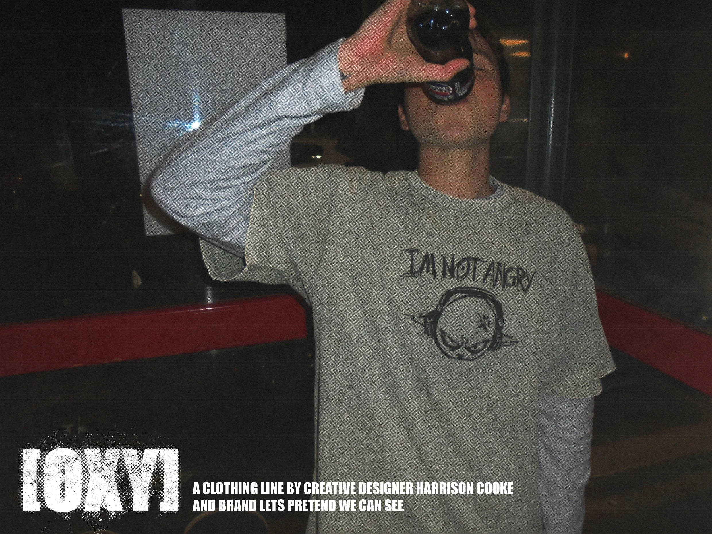Person holding a dark beverage bottle to their mouth. They are wearing a light grey sweatshirt with text and graphic on it. The background is dark with partially visible signage or posters, including part of the word 'TOXIC' and some text about cloth