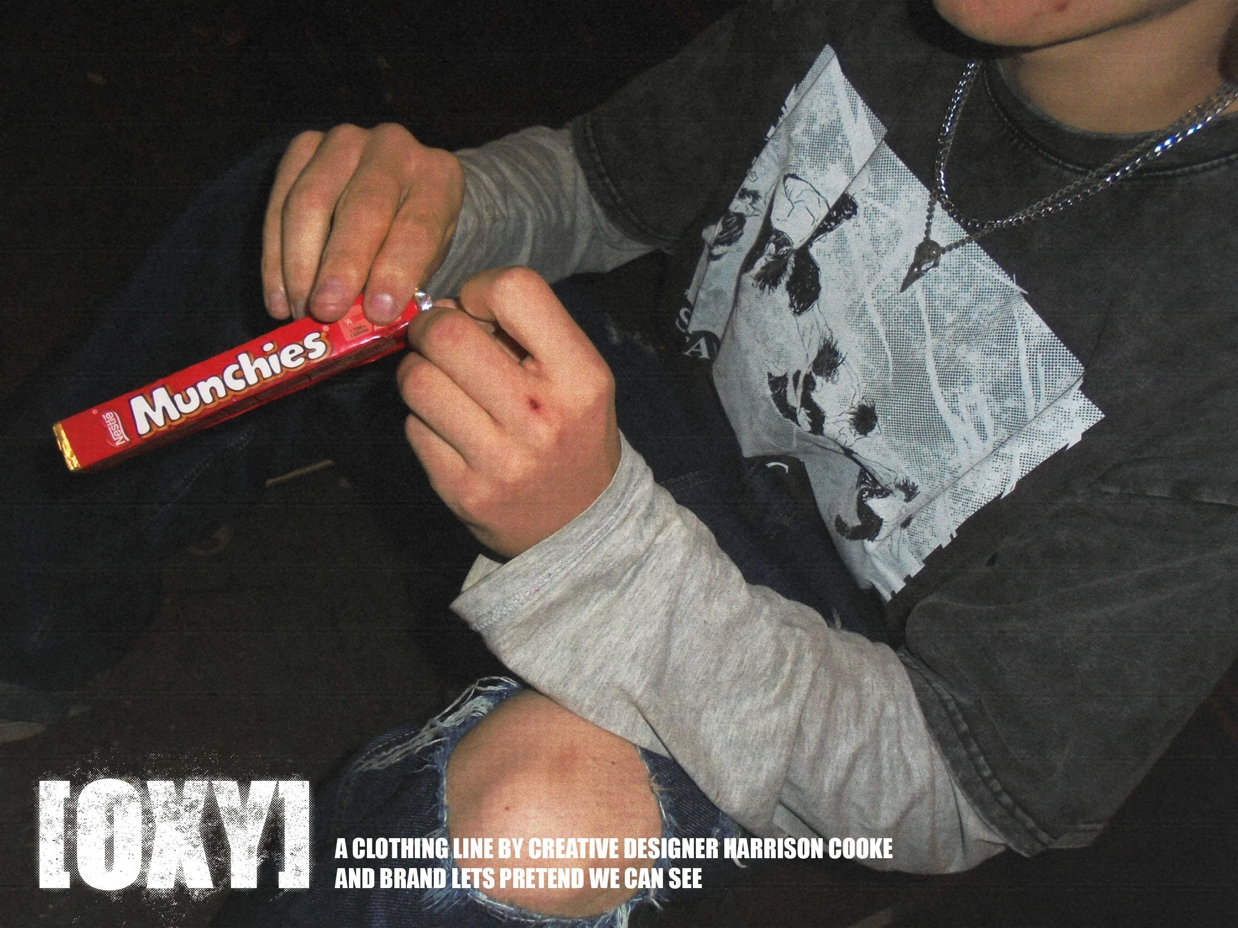 A person wearing a black and gray shirt with graphic designs and layered necklaces, holding a red Munchies snack bar, seated with another person's leg visible in the foreground.