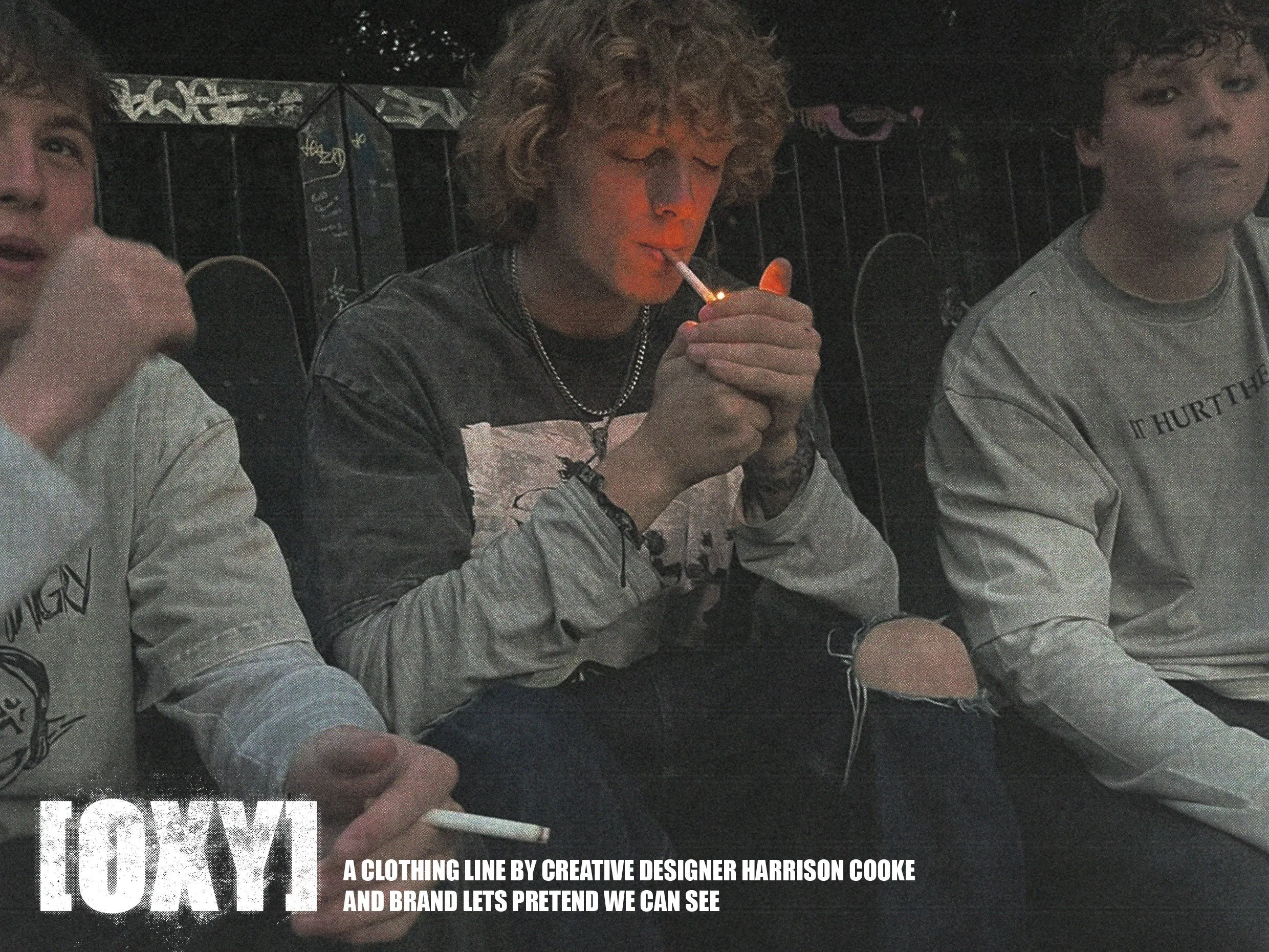 Three young men sitting together outdoors in low light, with the middle person lighting a cigarette. All are wearing casual clothing, including sweatshirts and jackets, and are positioned in front of a graffiti-covered wall.