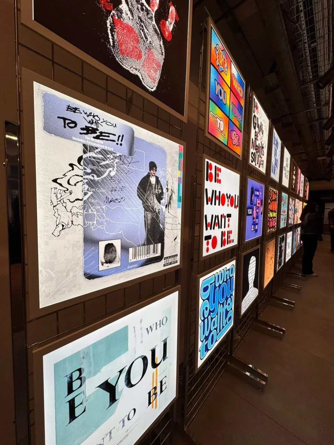 A wall display featuring illuminated posters with various artistic messages and designs, including motivational phrases, abstract art, and graphic prints, arranged in a grid formation in an indoor gallery or exhibition space.