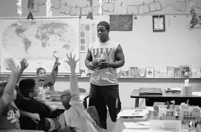 A teacher standing in front of a classroom of elementary students, holding a small ball, with students raising their hands.