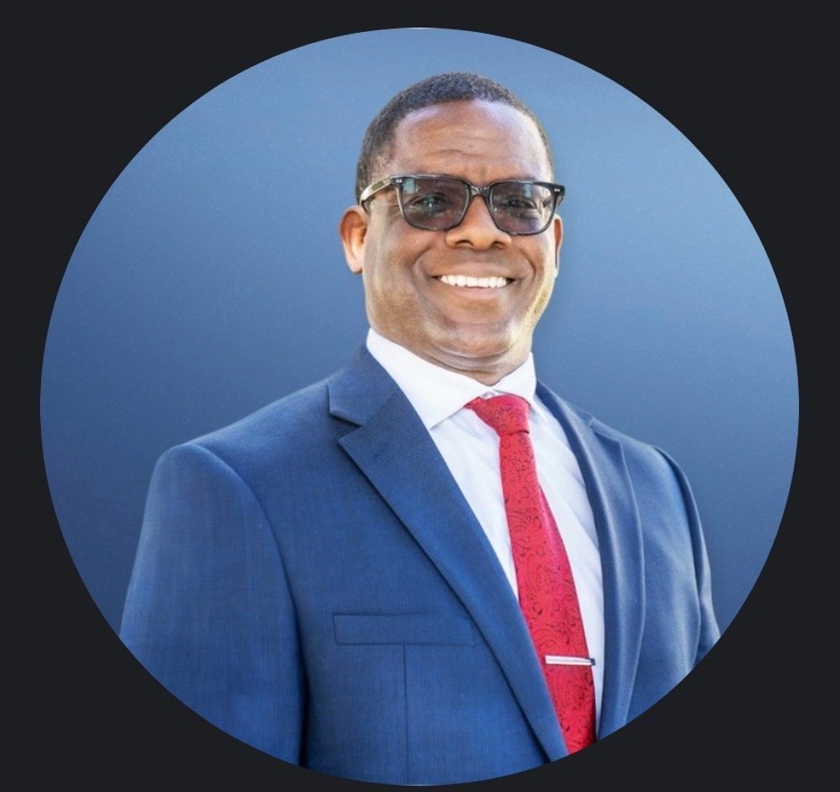 A smiling man wearing a dark blue suit, white shirt, red tie, and glasses, posing against a blue background.