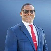 Smiling man in a blue suit with a white shirt, red tie, and sunglasses, standing outdoors against a clear blue sky.