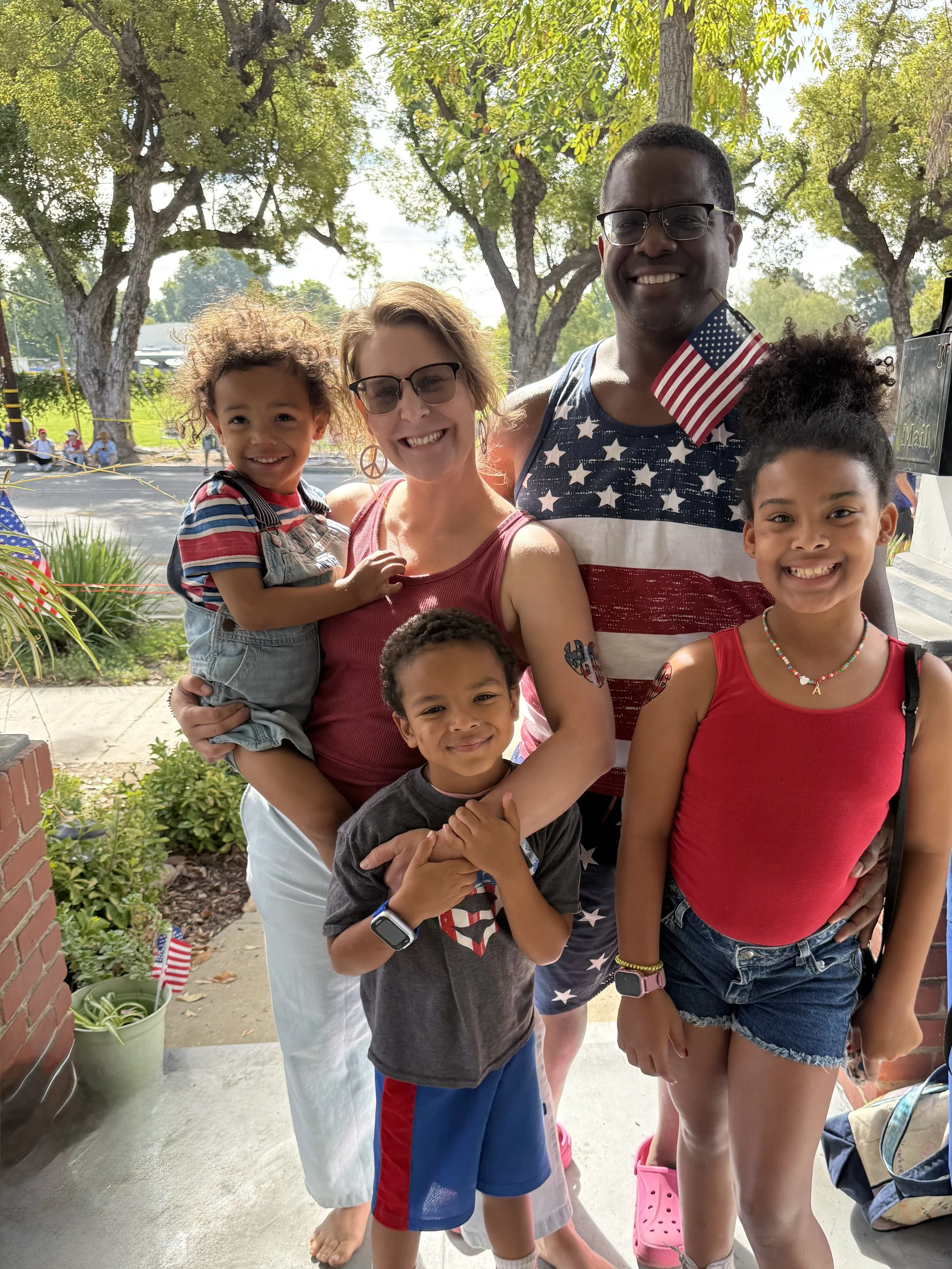 A group of smiling people posing outdoors, some holding small U.S. flags, in a lush, green area with trees and sunlight.