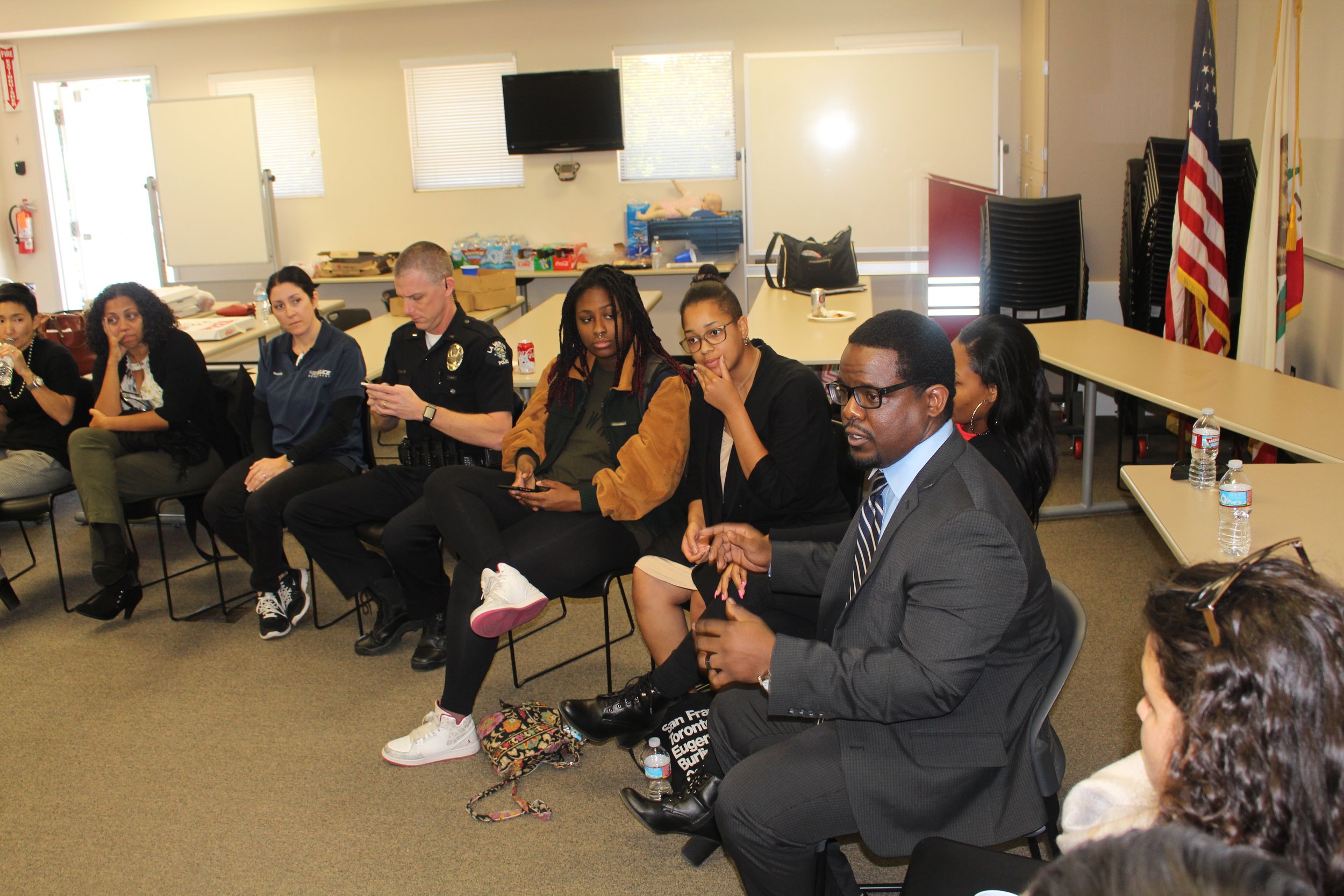 A diverse group of people sitting in chairs in a conference room with American flags and stacked chairs in the background.