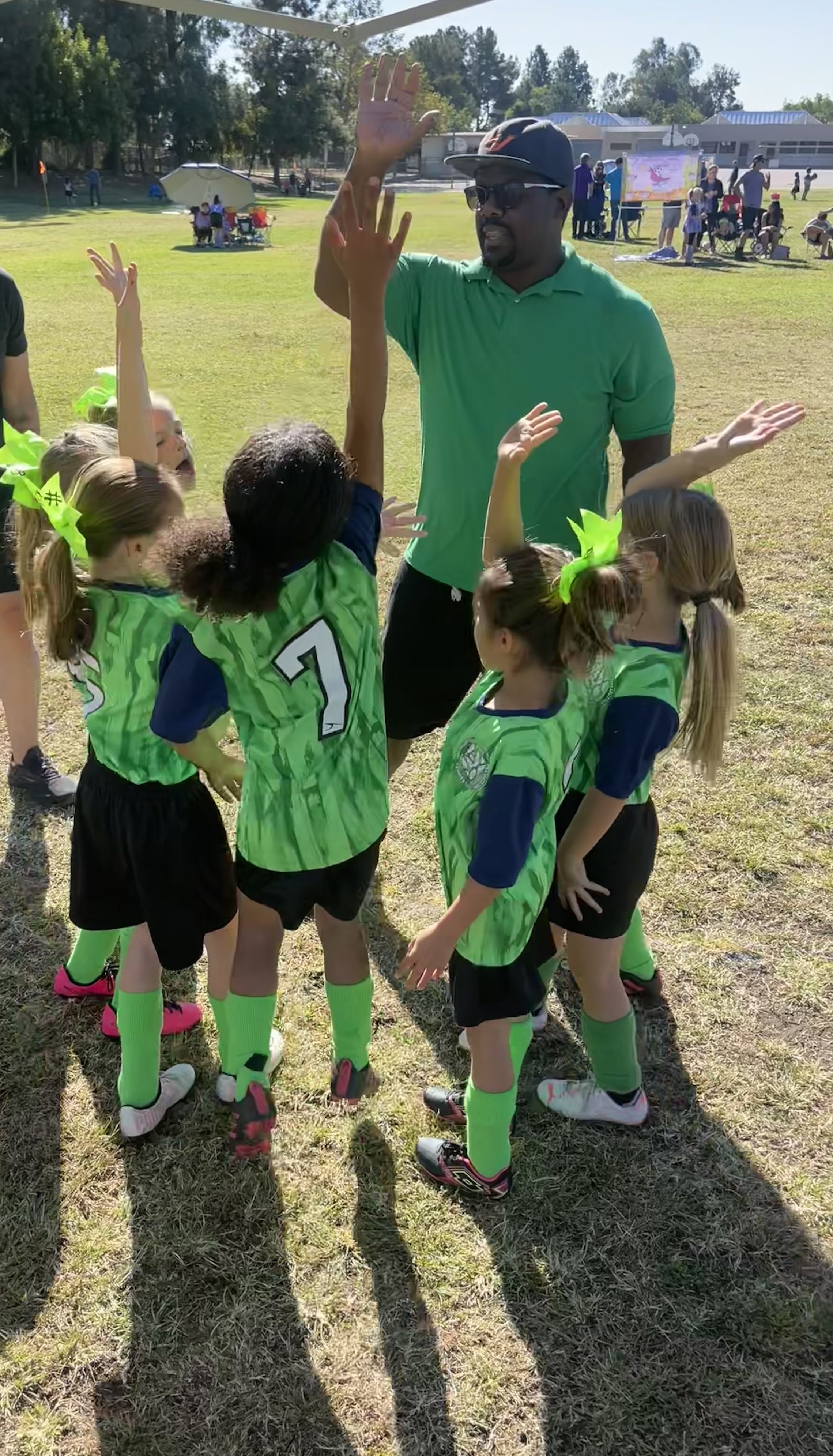 A man in a green polo shirt and black shorts giving a high-five to a group of young girls in matching green sports uniforms with neon green socks outdoors on a sunny day, with other children and adults in the background.