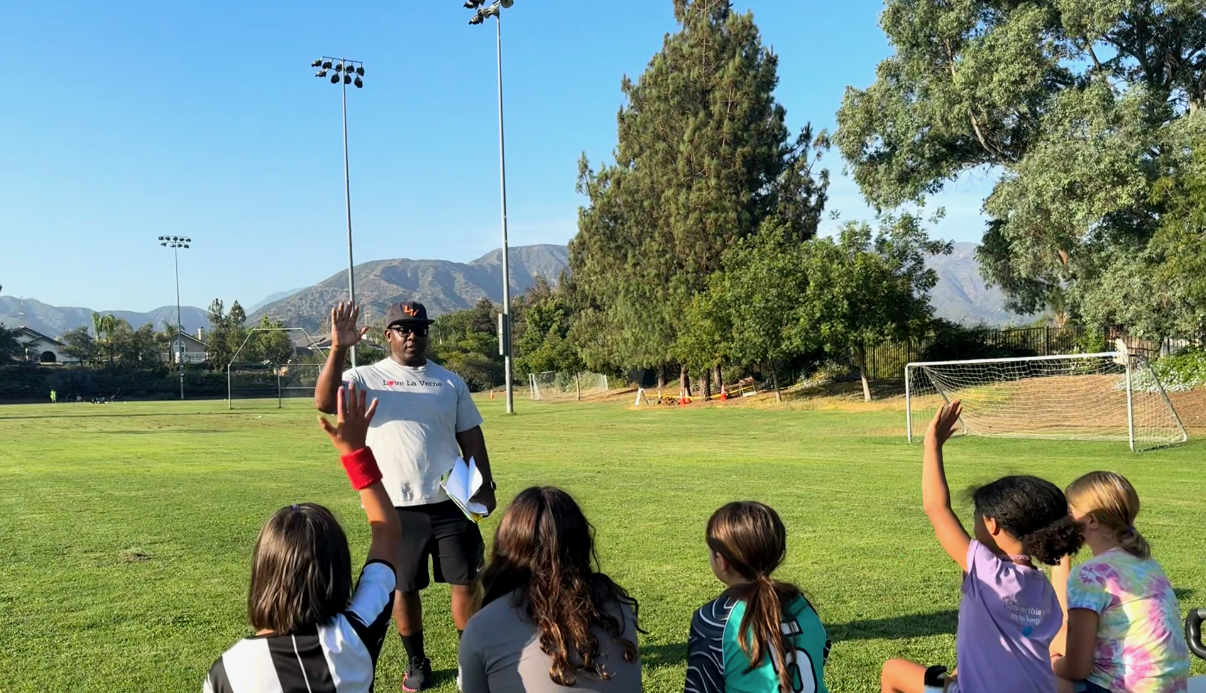 Coach giving instructions to a girls' soccer team on the field, with some players raising their hands.