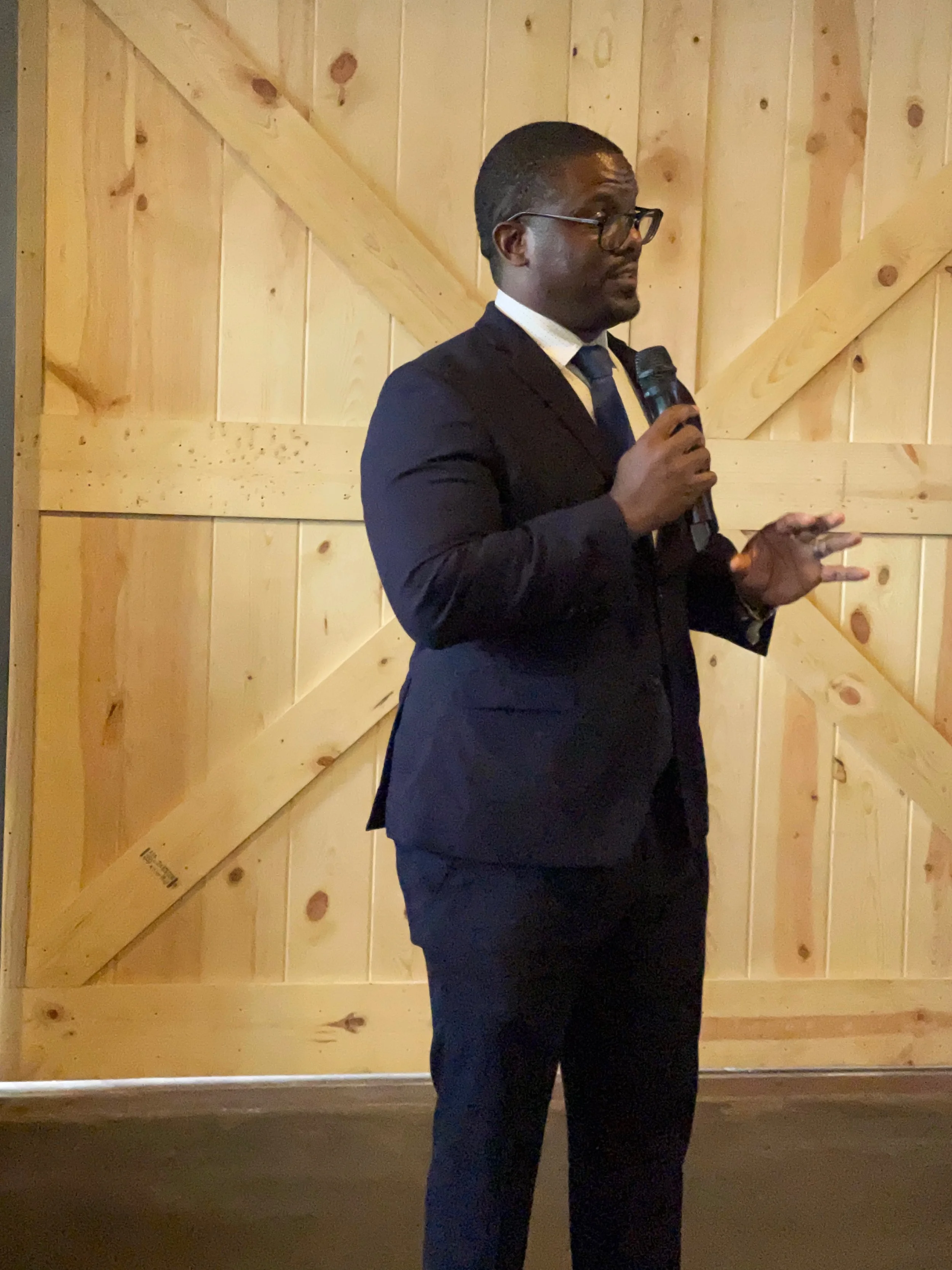 An African American man in a dark suit, glasses, and a tie standing in front of a light wooden wall, holding a microphone and gesturing with his other hand, likely giving a speech or presentation.