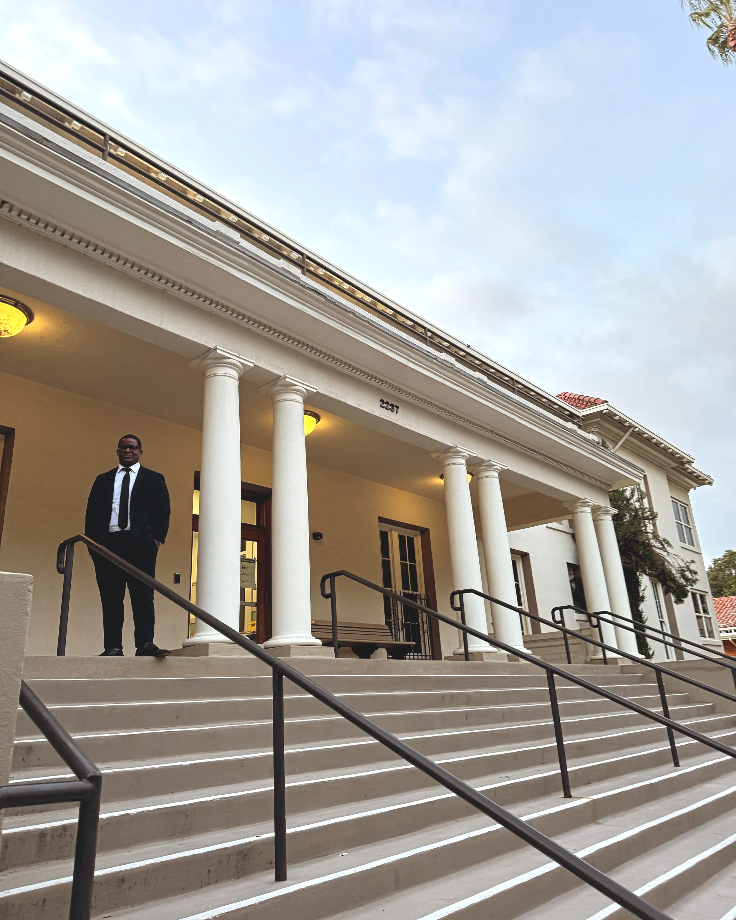 A man in a suit standing at the bottom of a set of steps leading up to a building with white columns and a red-tiled roof. The building has lights under the overhang and the sky in the background is partly cloudy.